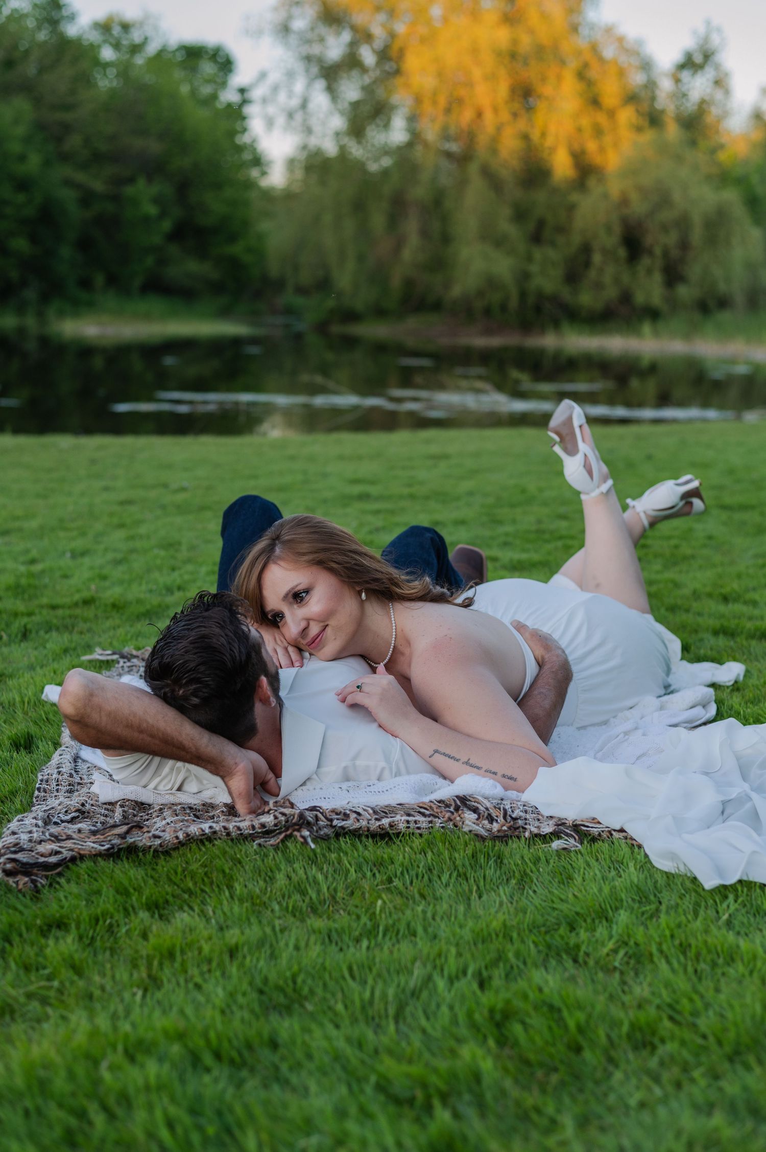 Couple lying on a blanket in lush green park near a pond, woman in white dress smiling at camera.