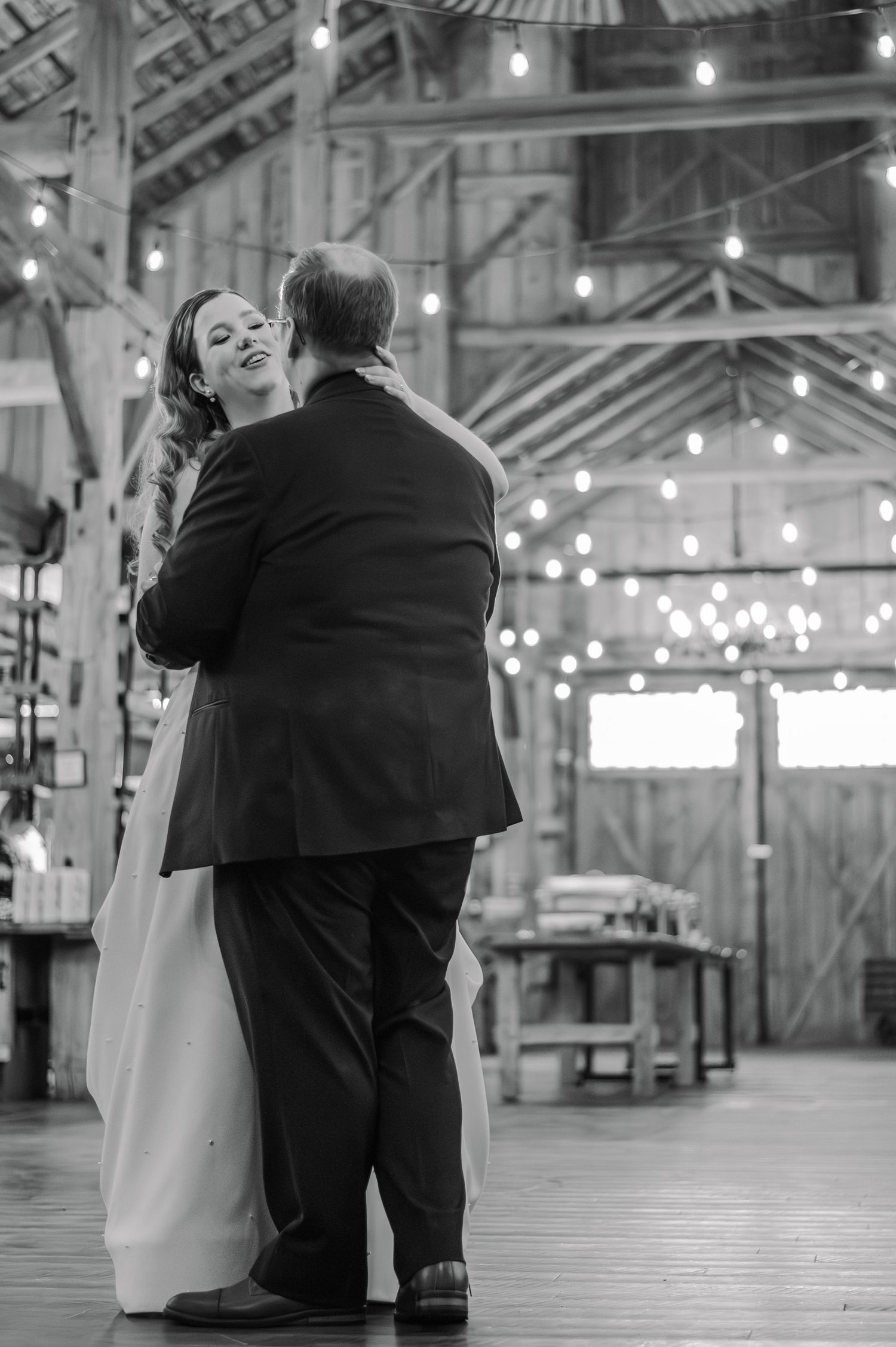 A black and white wedding photo series showing a couple dancing intimately in the rustic Brussels Four Winds Barn with string lights.