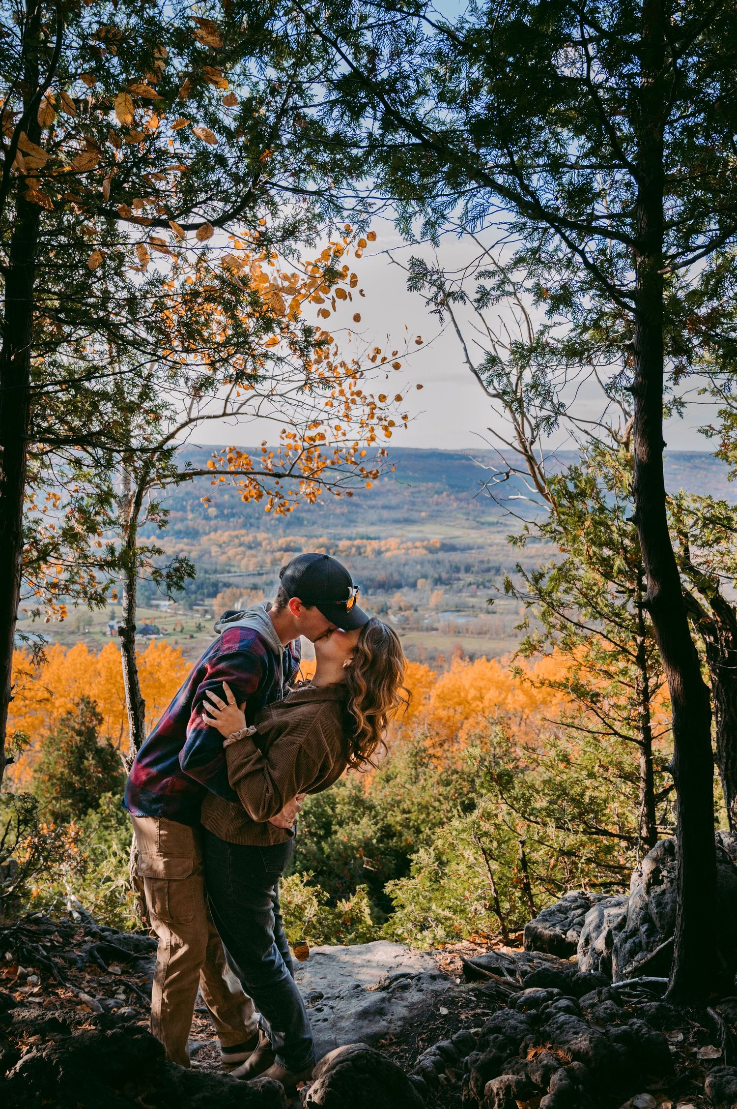 A couple shares a romantic moment on a mountain overlook during autumn with colorful fall foliage in the valley below.