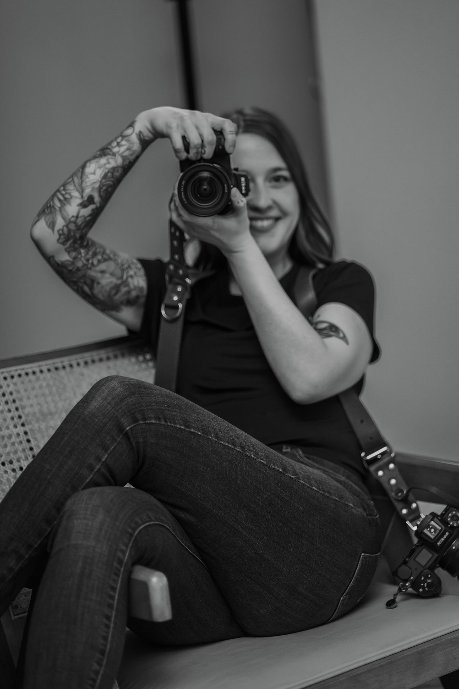 Black and white photo of a tattooed woman smiling while holding a DSLR camera, seated casually in jeans and a black t-shirt.