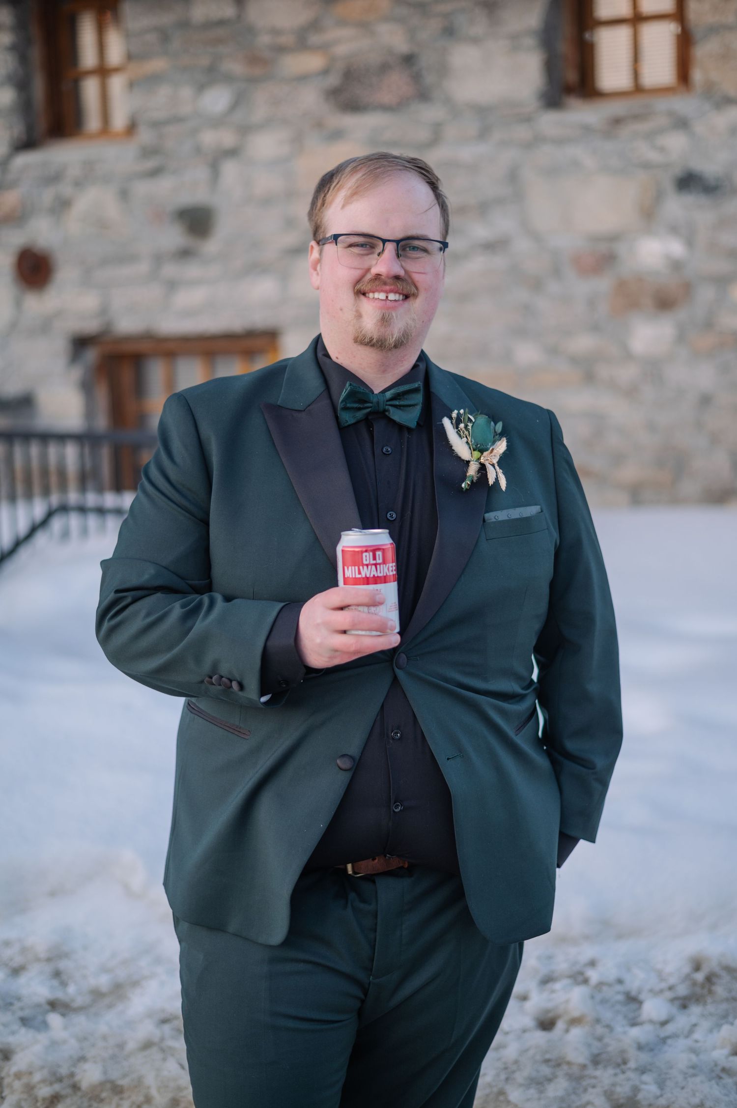 Groom in green tuxedo with bow tie and boutonniere holds a beer can, posing outdoors in winter snow.