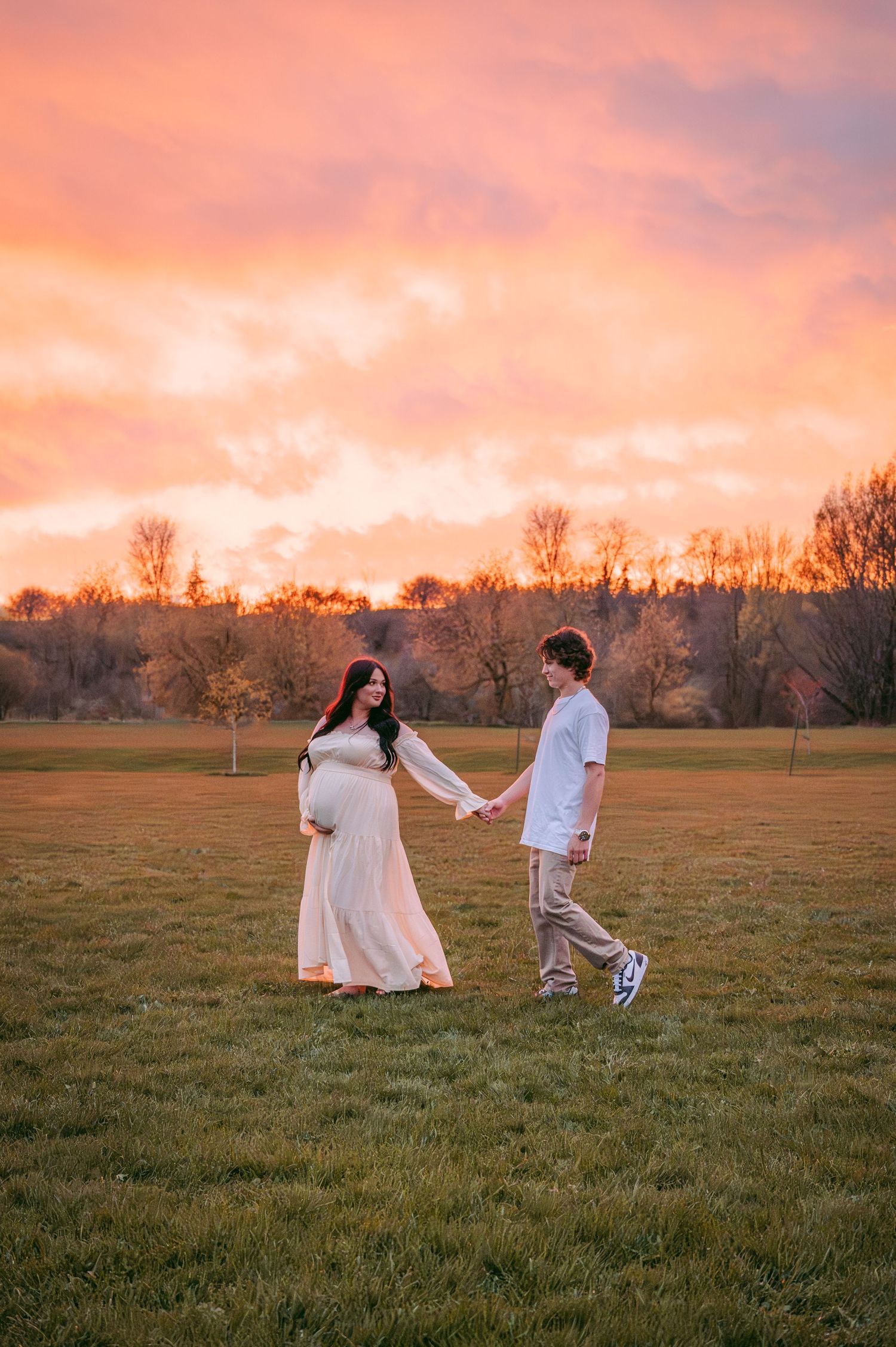 Couple holds hands during golden hour maternity shoot in an open field.