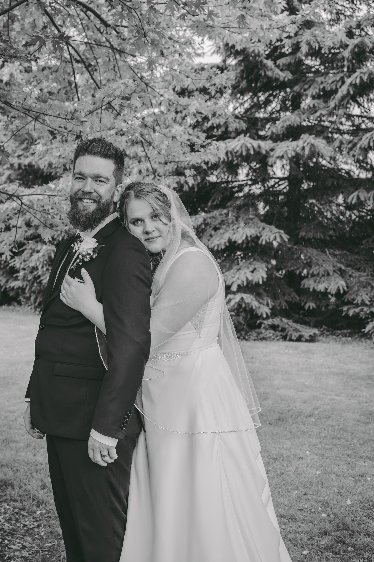 Black and white portrait of newlyweds embracing in front of evergreen trees.