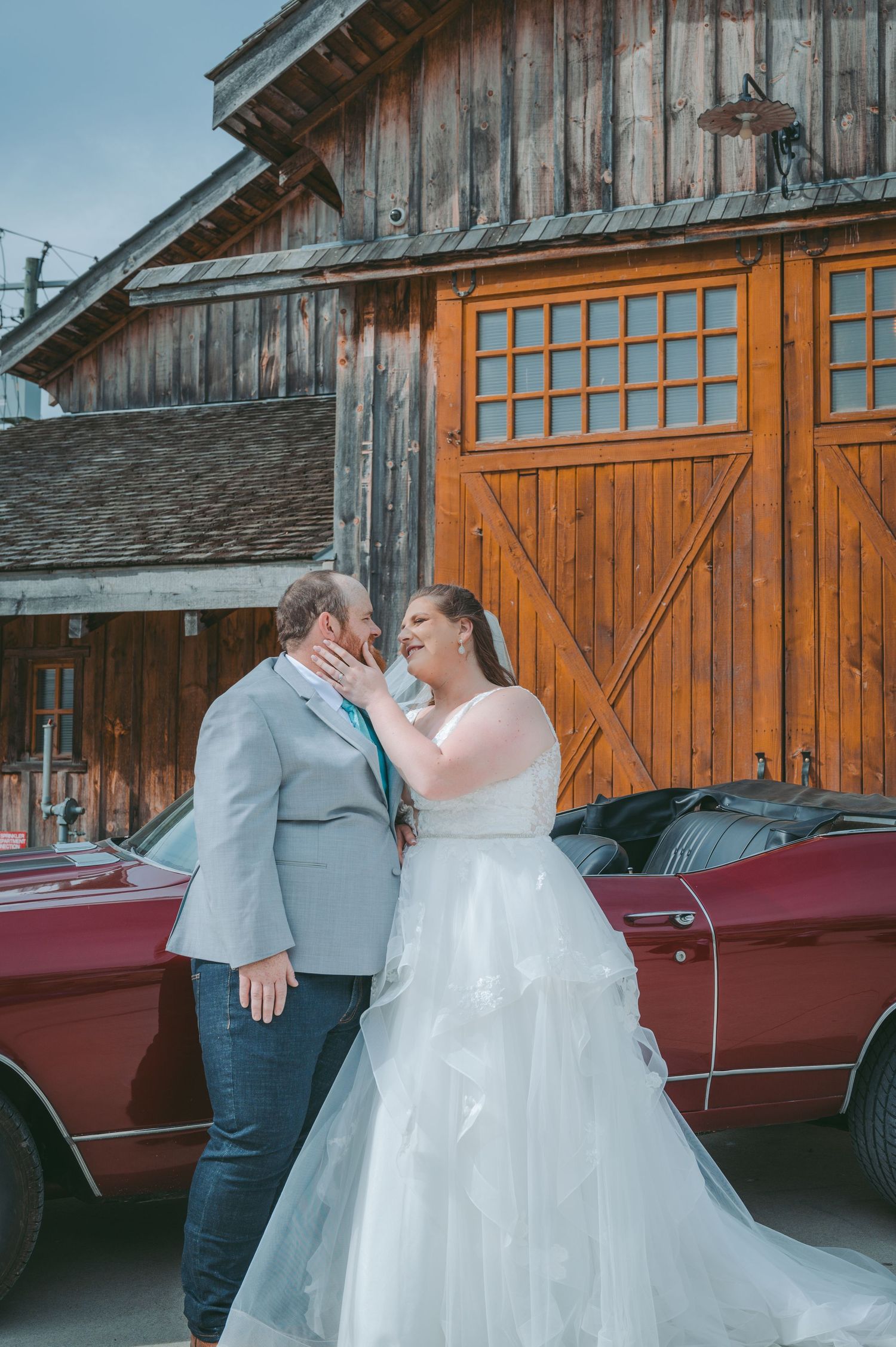 A bride and groom share an intimate moment outside a rustic barn with orange doors and a vintage red car.
