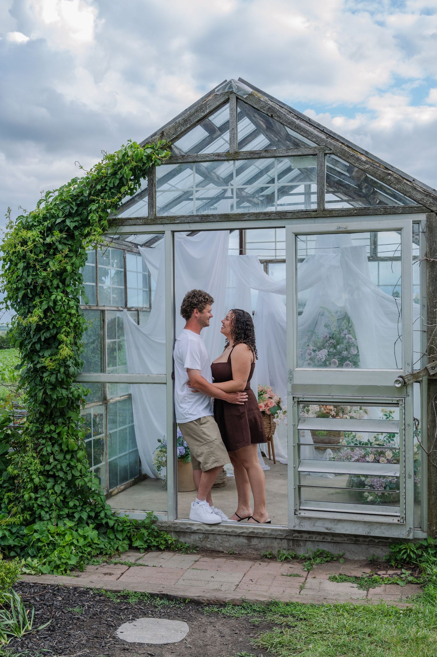 Couple embracing inside a charming glass greenhouse with ivy, white curtains, and a bouquet on a cloudy day.