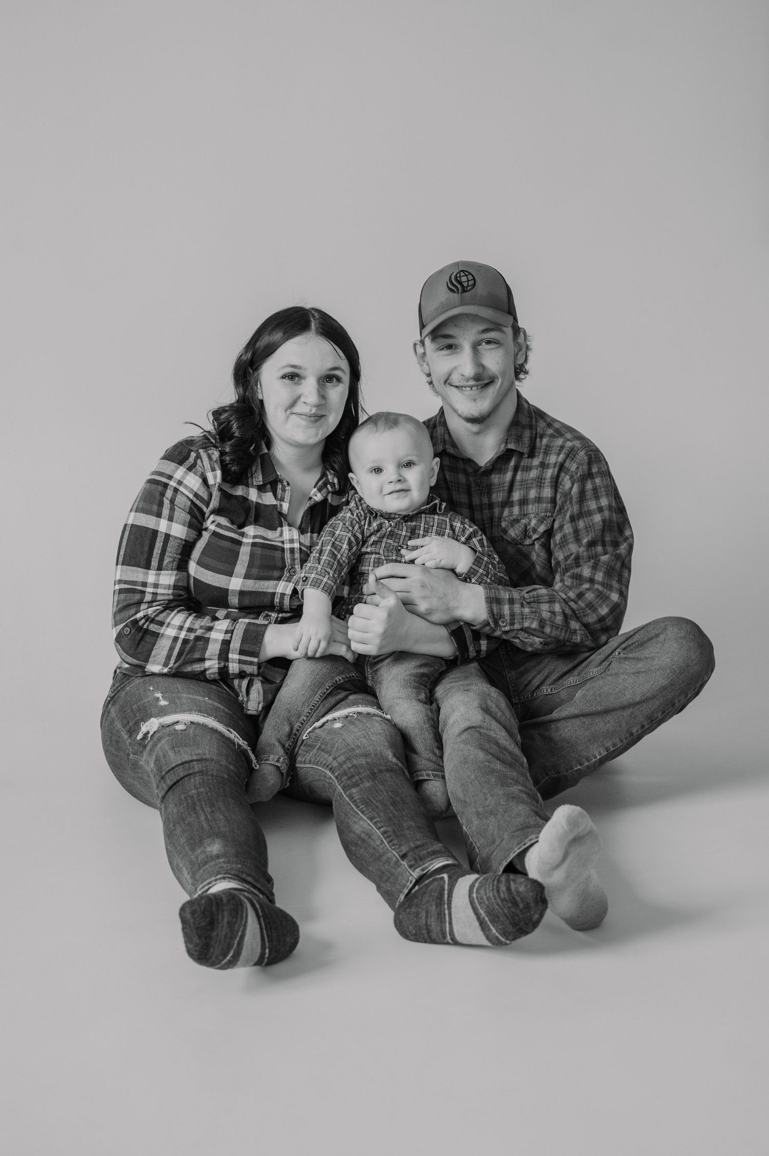 Black and white family portrait of two parents in flannel shirts sitting on floor holding their smiling baby.