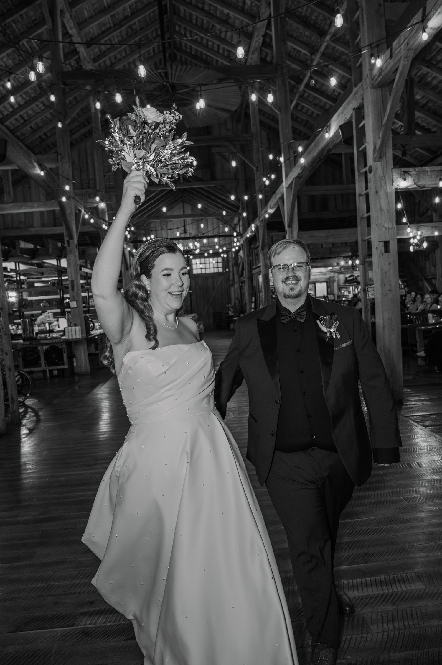 Black and white photo of a joyful bride raising bouquet and groom in tuxedo walking through a rustic barn wedding venue.