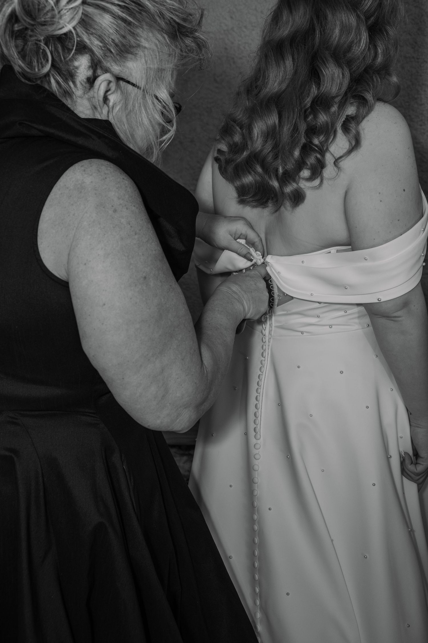 Black and white photo of a woman fastening the back of a bride's white wedding dress before the ceremony.