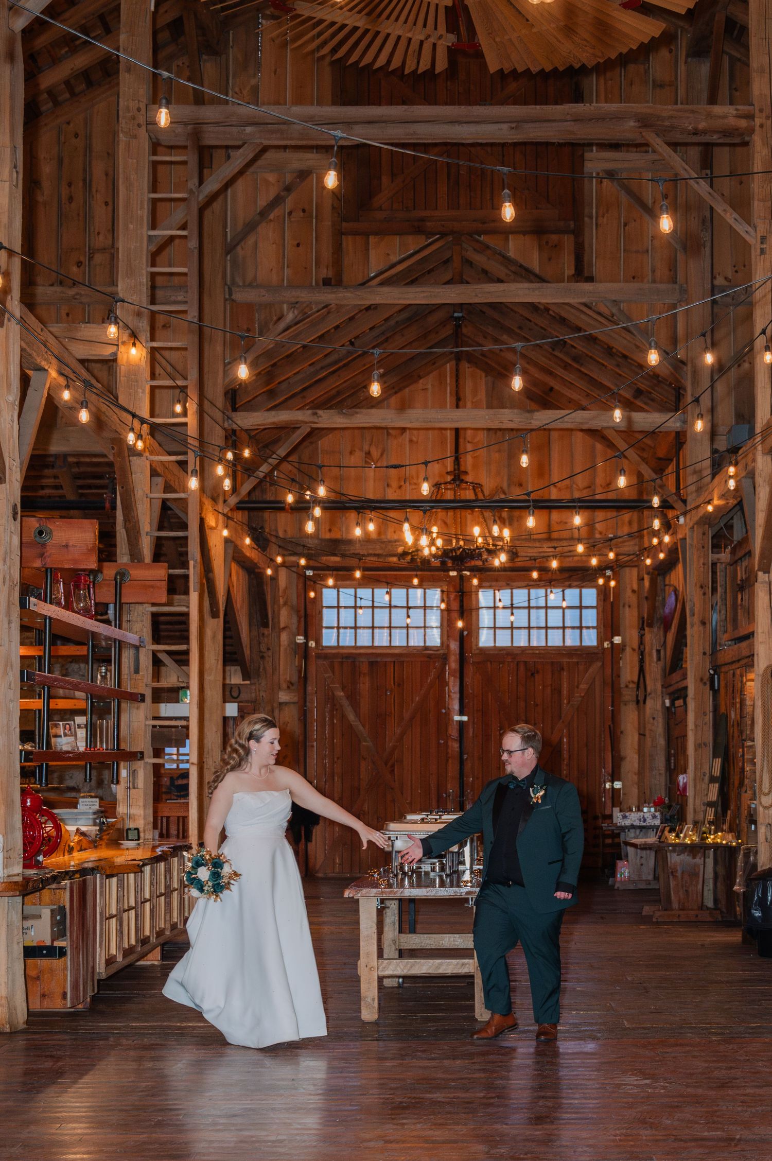 Bride and groom hold hands in a rustic barn wedding venue adorned with warm string lights and wooden beams.