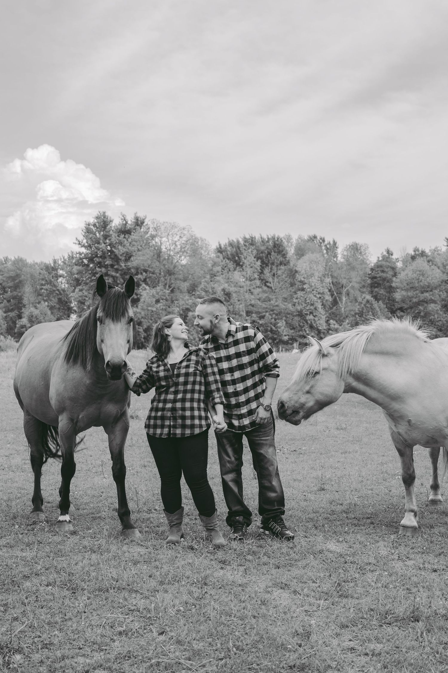 A couple stands between two horses in a black and white countryside photo.