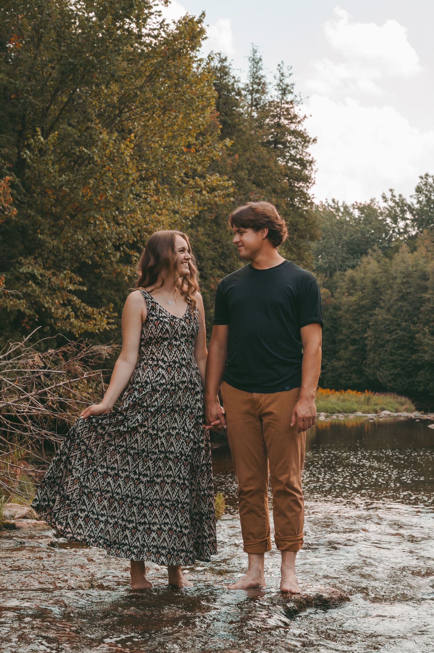 Couple holding hands while standing in river water with autumn trees in background.