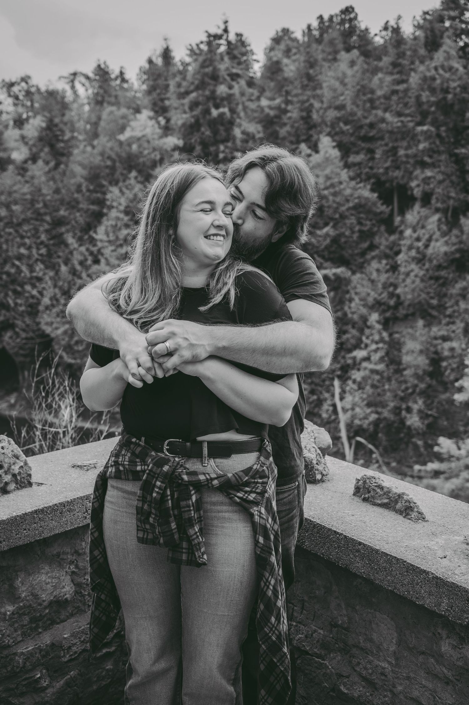 Intimate black and white portrait of couple embracing outdoors with forest backdrop.