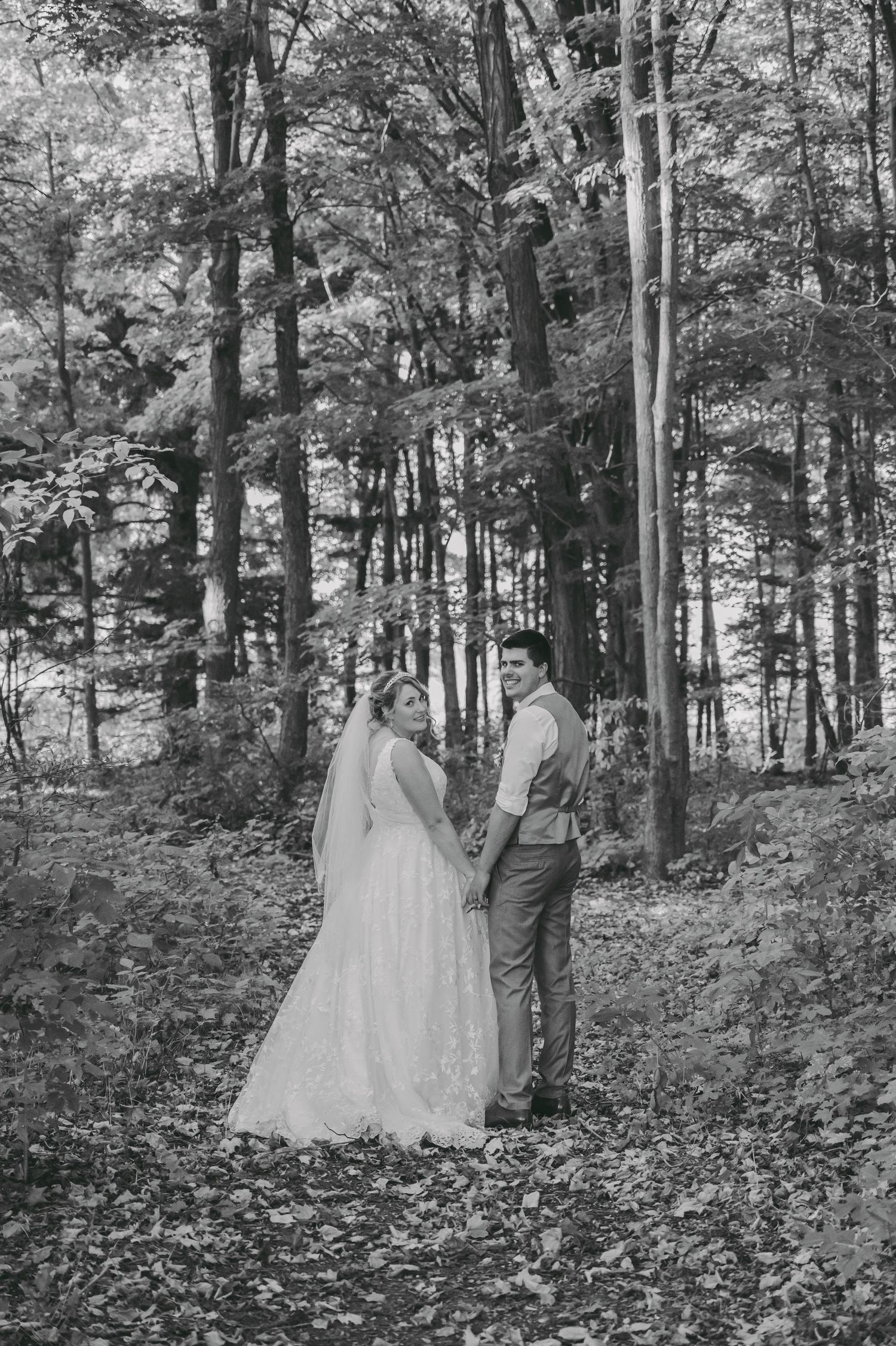 Black and white portrait of couple in wedding attire standing together in wooded forest setting.