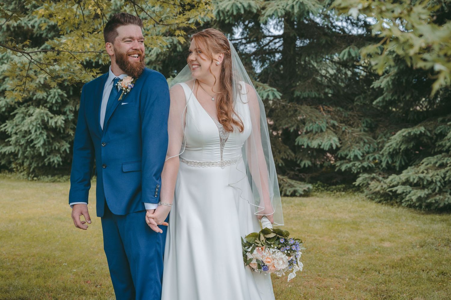 A couple in wedding attire walks together and laughs in a garden setting with green trees in the background.