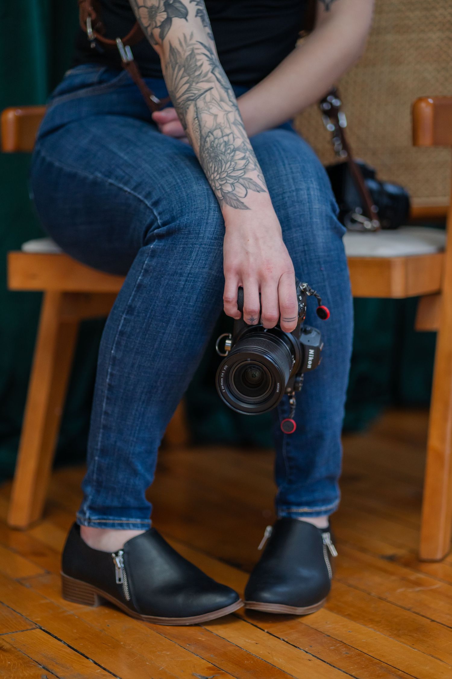 Photographer in blue jeans and black ankle boots holds camera while sitting, showing detailed floral arm tattoo.