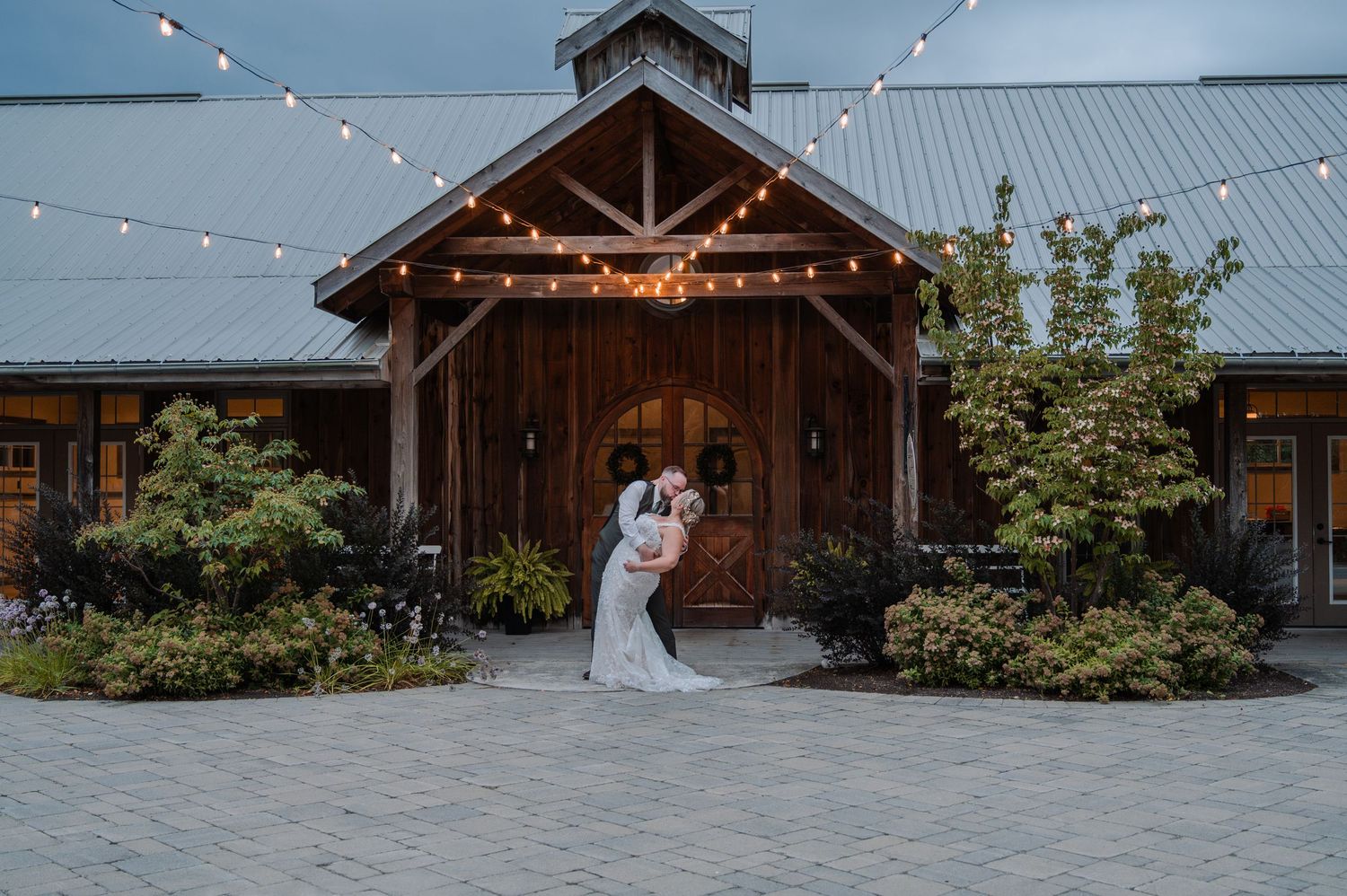 Couple sharing a romantic kiss outside a rustic barn wedding venue with string lights at dusk.