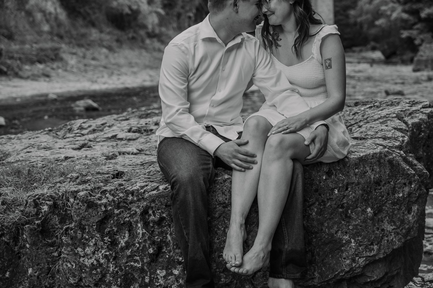 Two people sitting close together on rocky ledge during outdoor engagement photo session in black and white at the Elora Gorge.