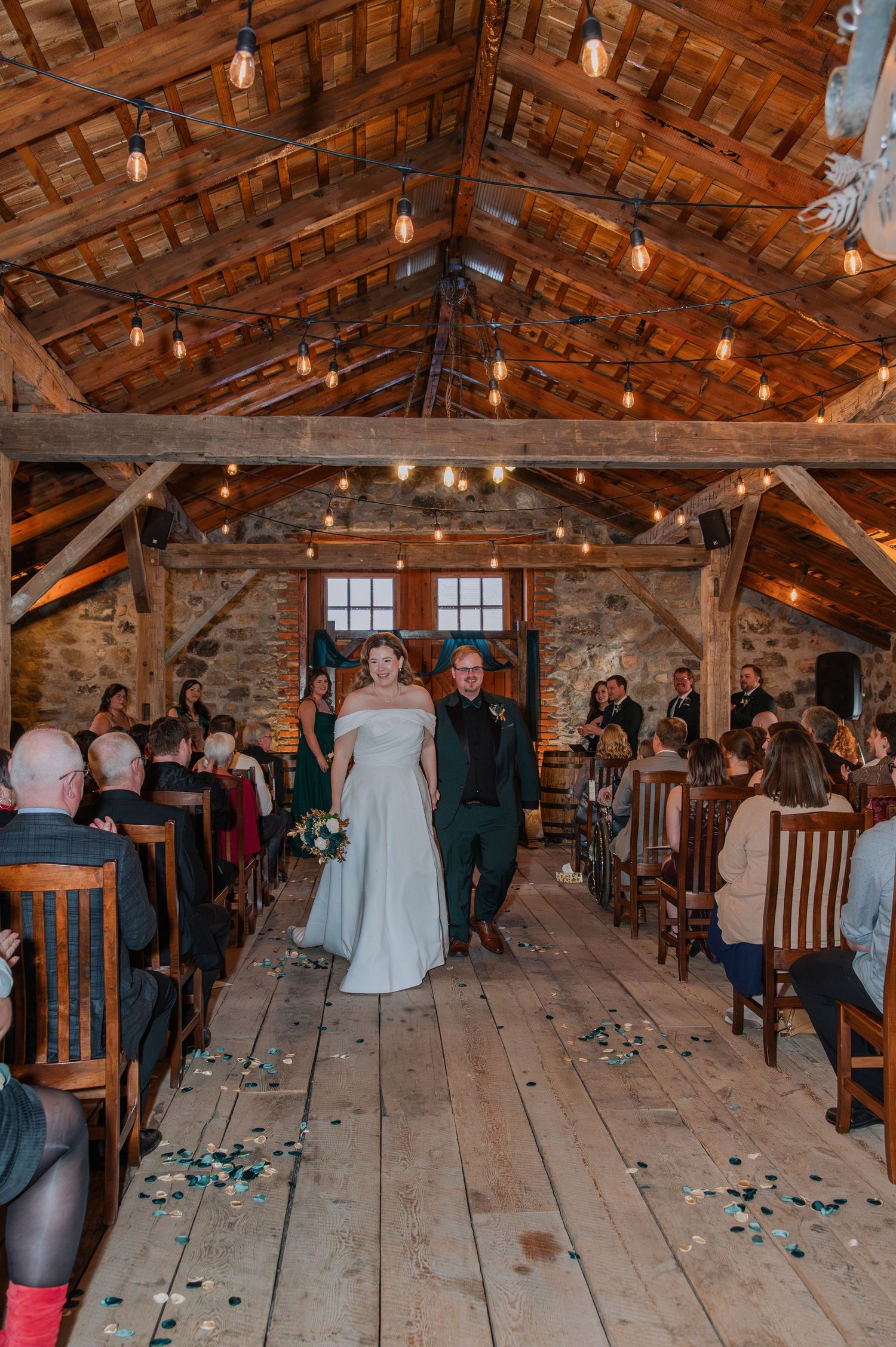 Bride and groom walk down the aisle of a rustic barn wedding venue with string lights and stone walls.