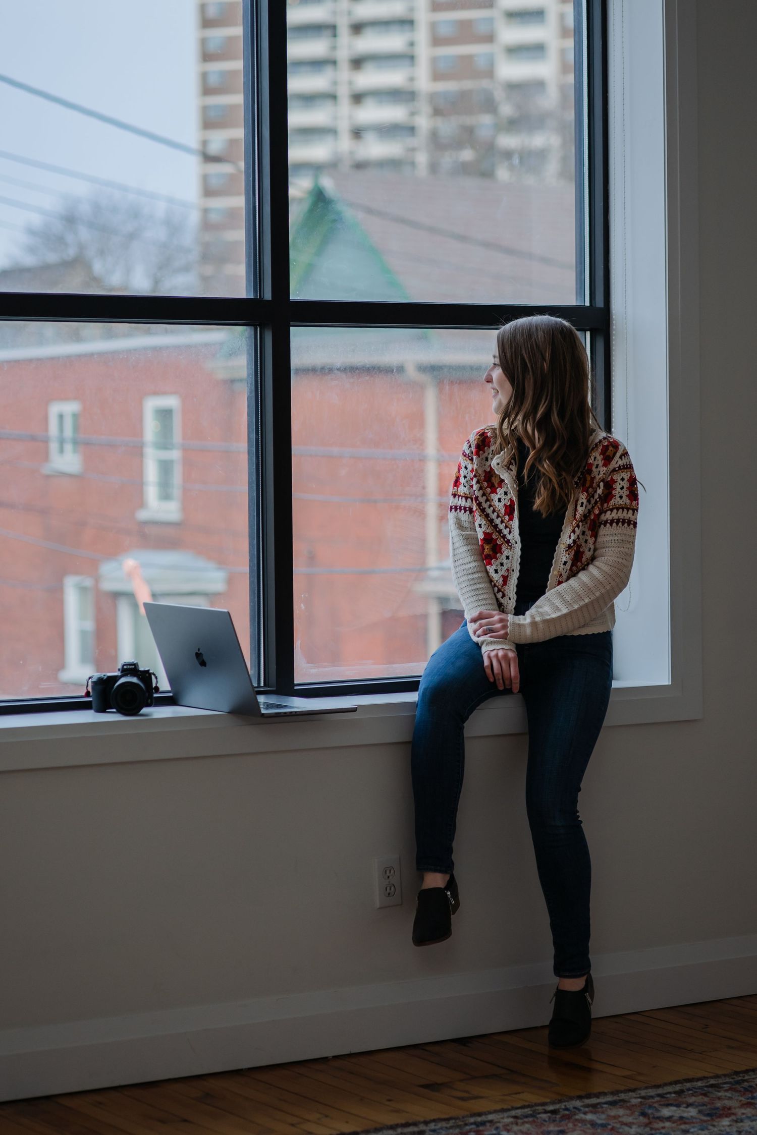 Person in cozy patterned sweater sits by window with laptop in urban apartment setting with brick buildings outside.
