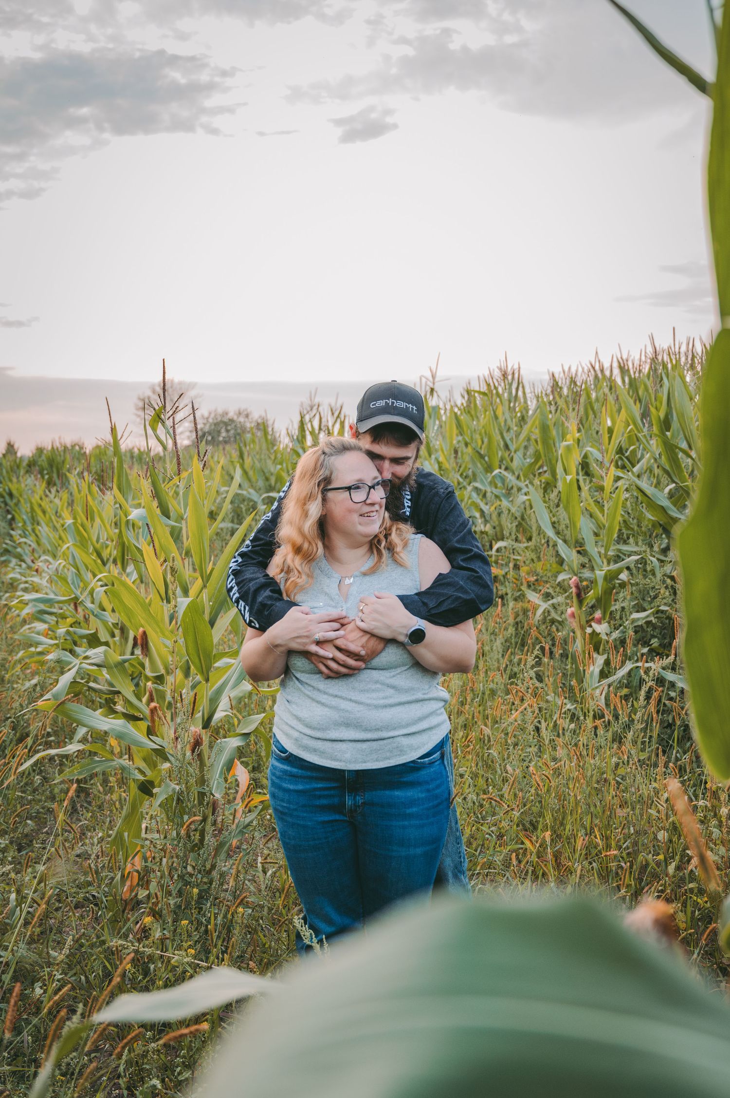 A couple embraces in a tall cornfield on a cloudy summer day.