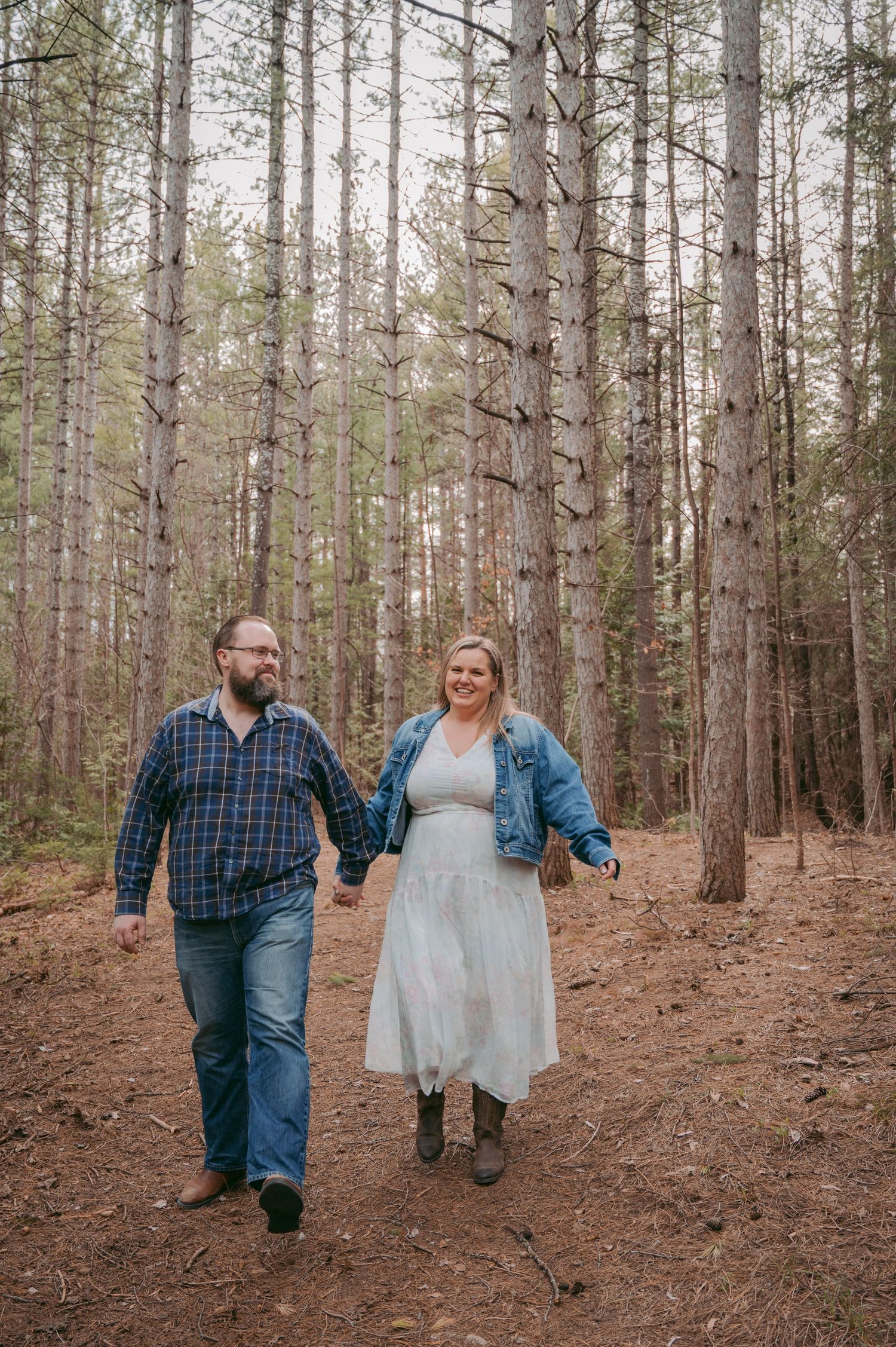 Couple walking hand in hand through a pine forest wearing casual attire and boots.