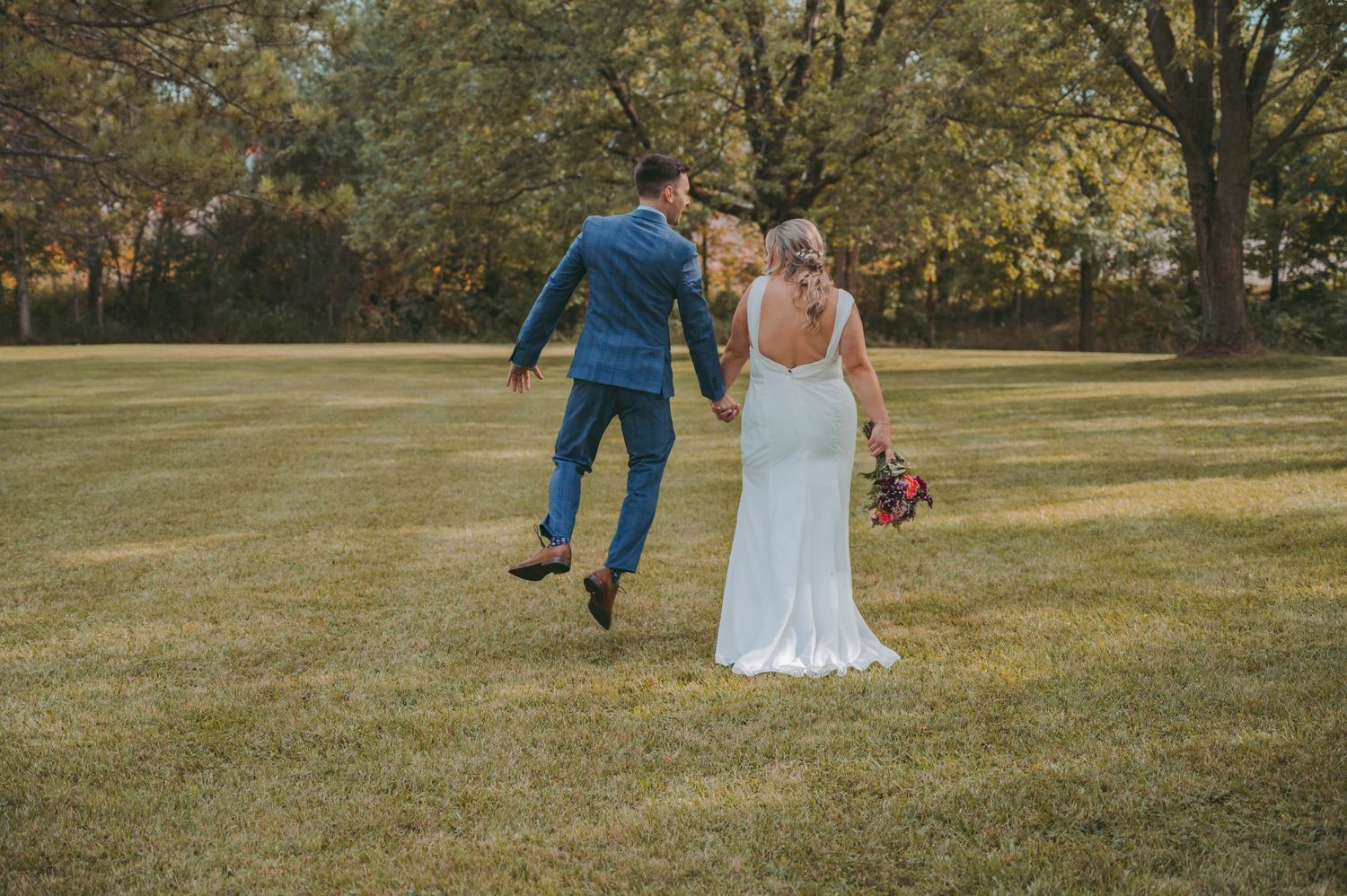Playful couple walks across grassy field during wedding photoshoot.
