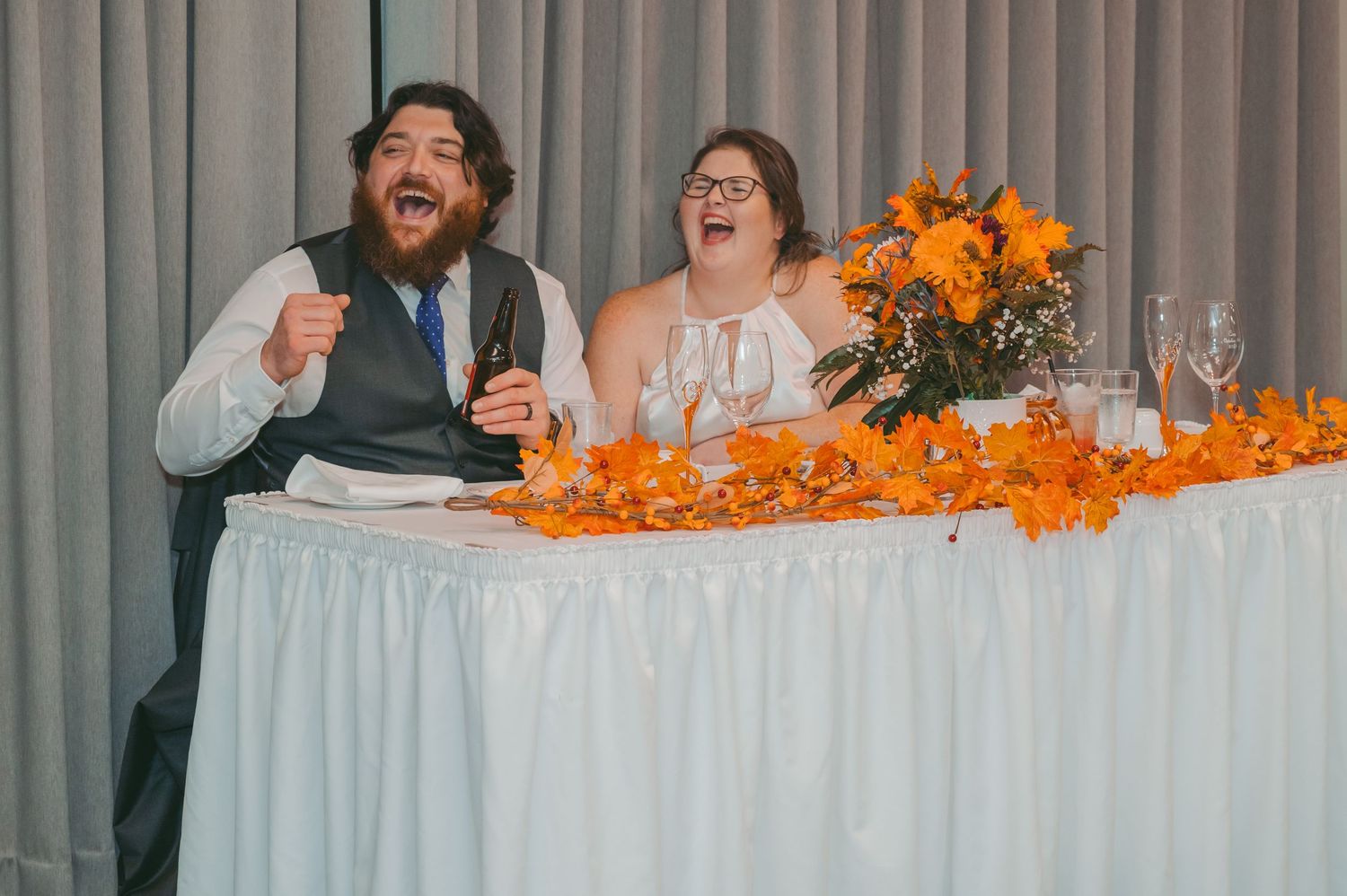 A joyful candid moment of newlyweds laughing at their reception table decorated with orange flowers.