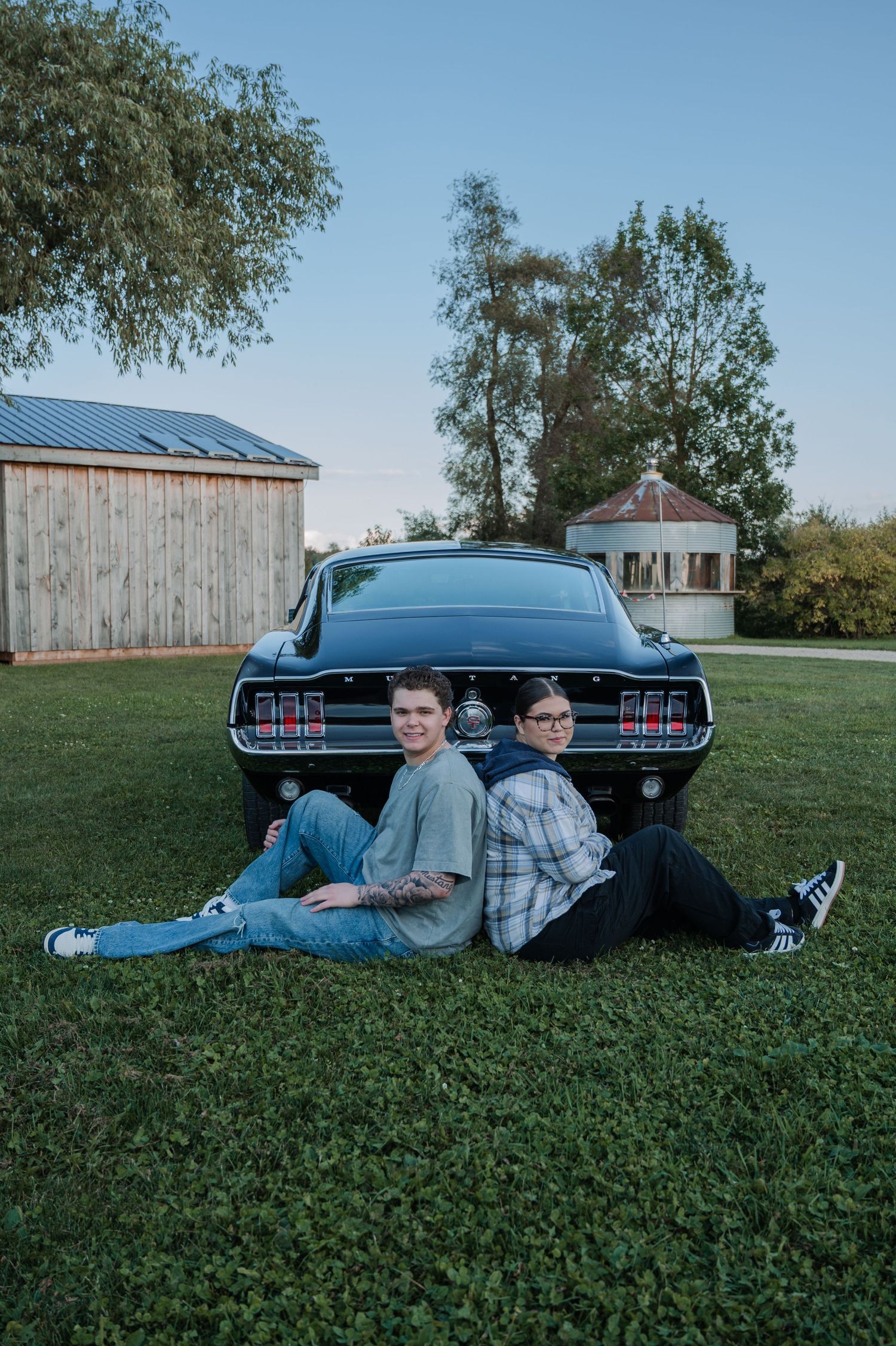 Two people lounging on grass next to a classic black muscle car with vintage taillights during a sunny day.