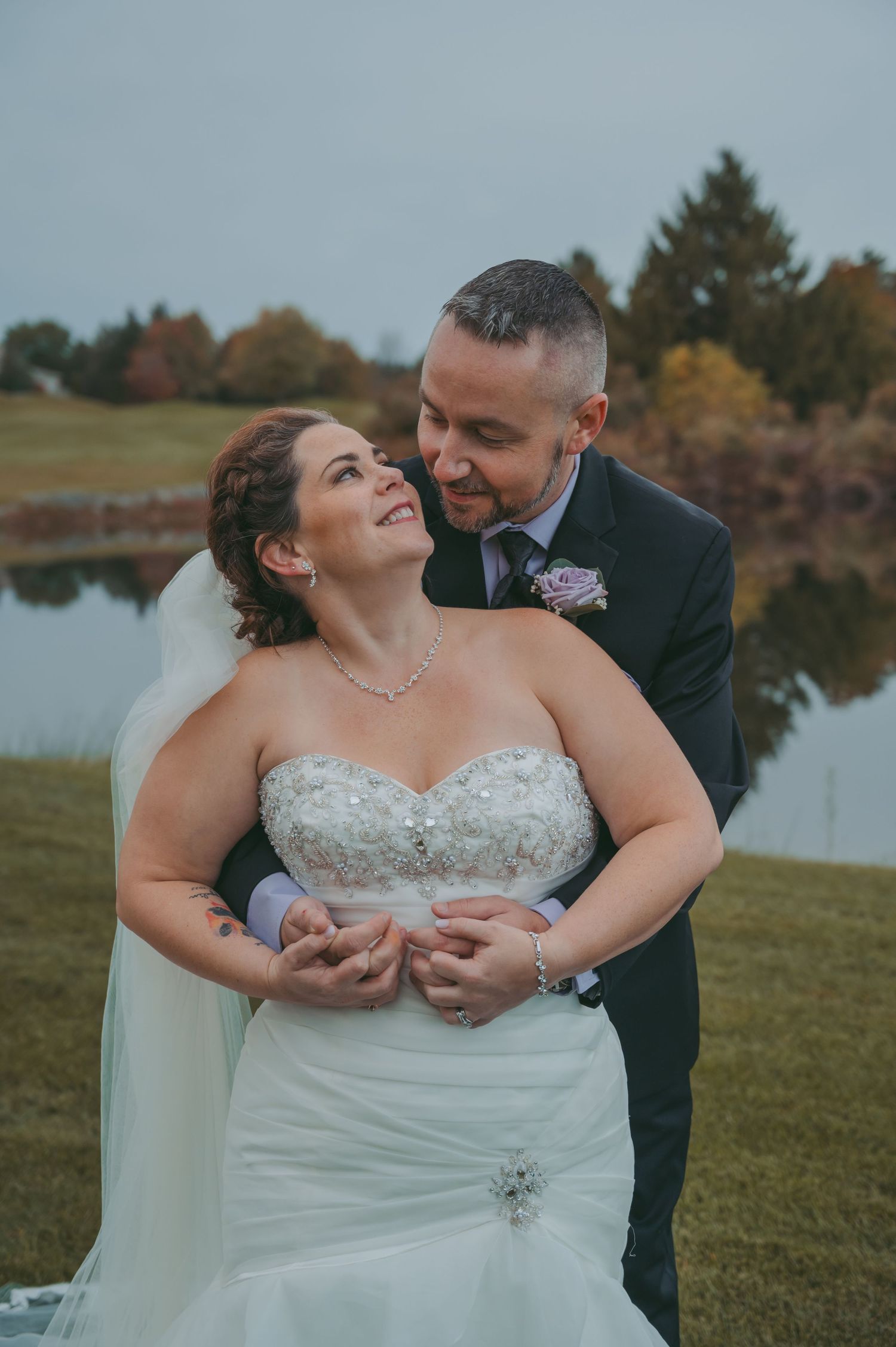 Tender moment between newlyweds by a reflective pond surrounded by autumn trees.