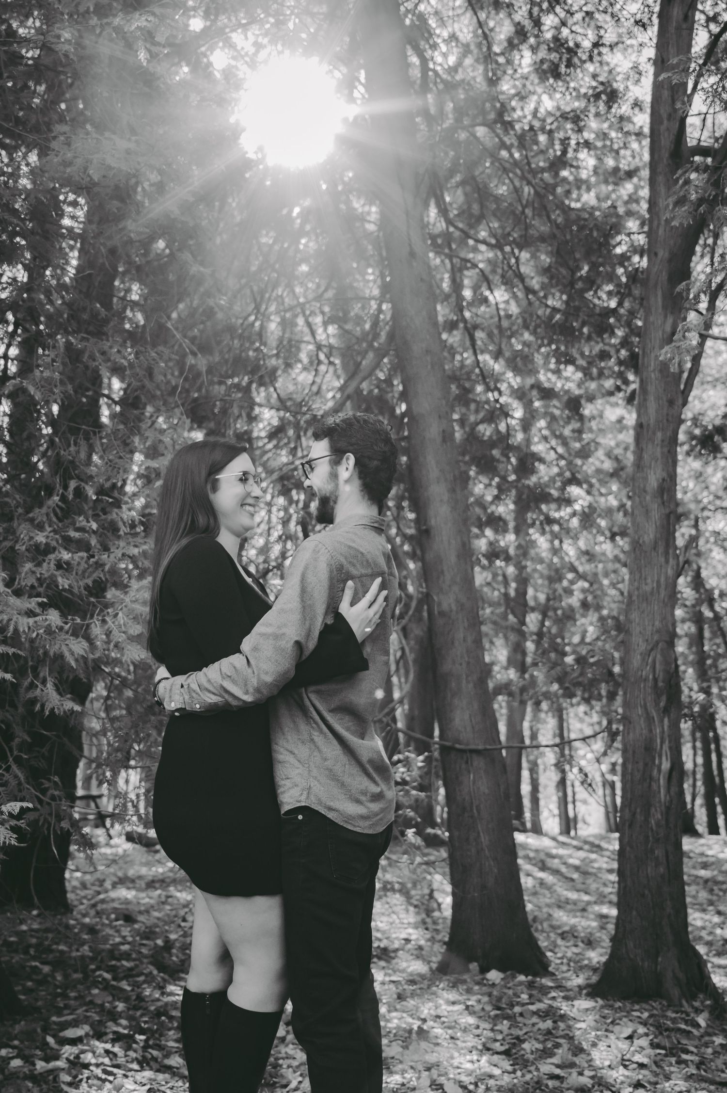 A romantic black and white photo of a couple holding each other in a sunlit forest setting.