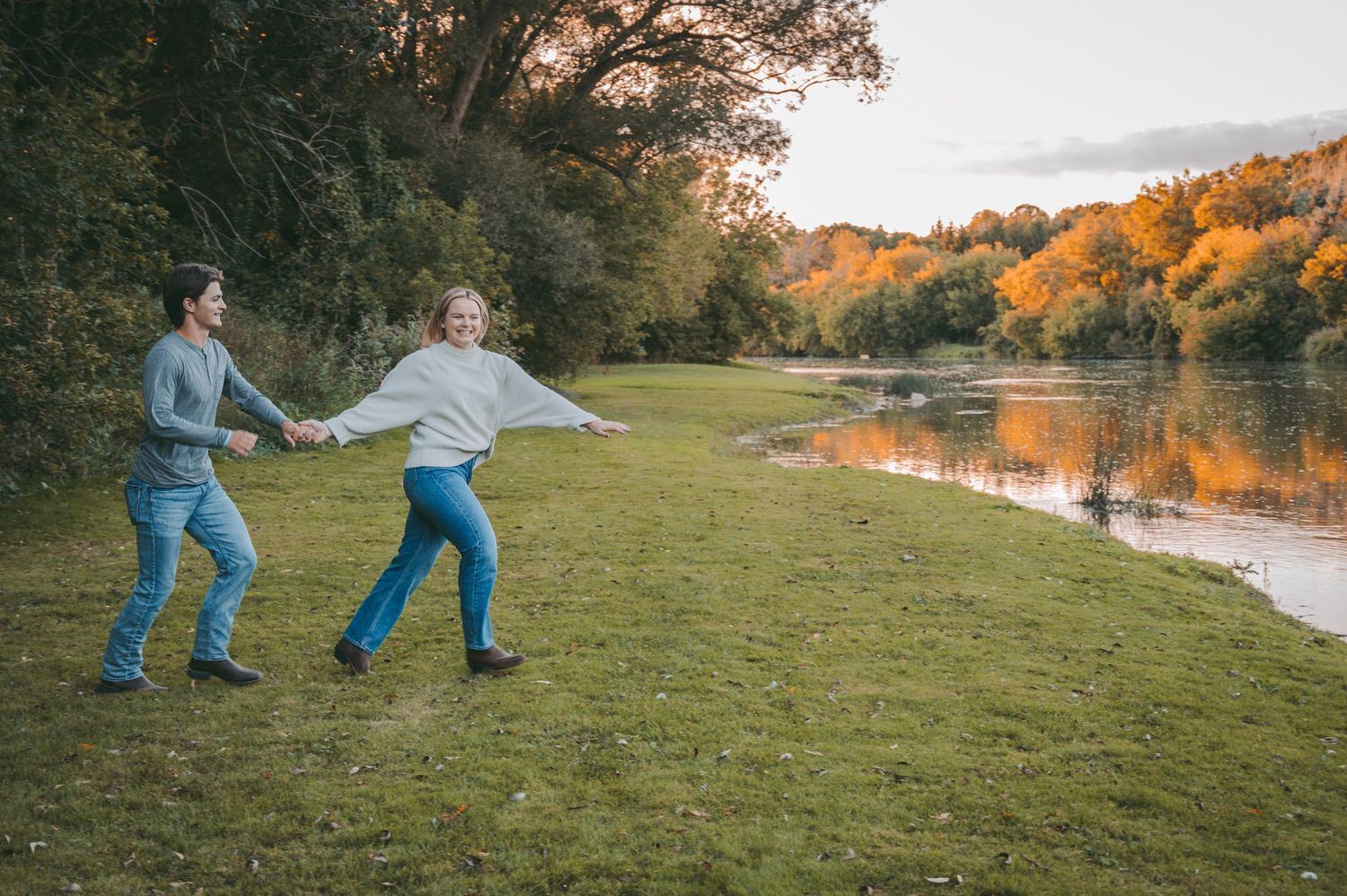 A playful couple dances together on grass near a lake with fall colors in the background.