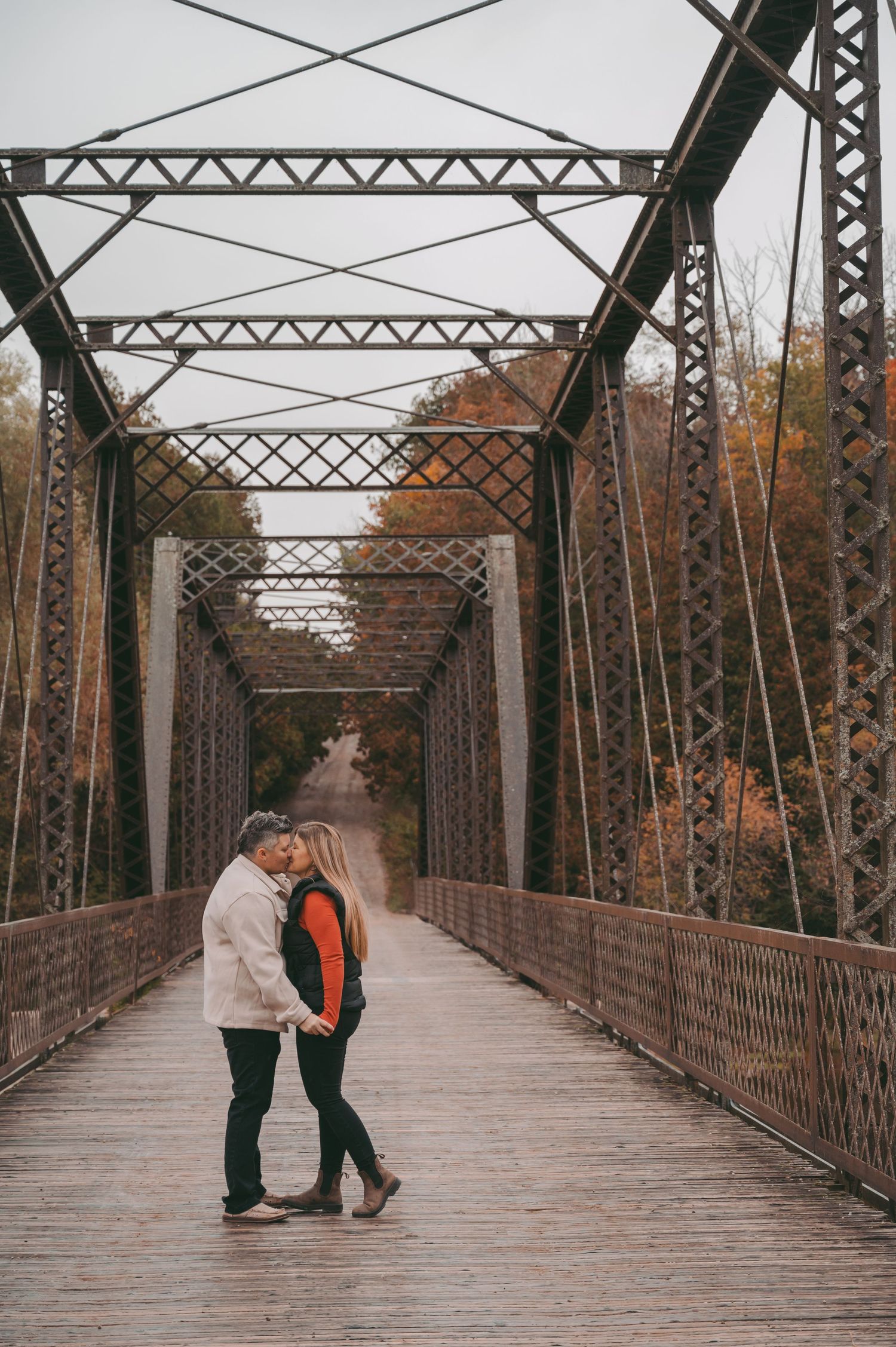 A couple embracing on a rustic wooden bridge surrounded by fall colors and metal trusses.