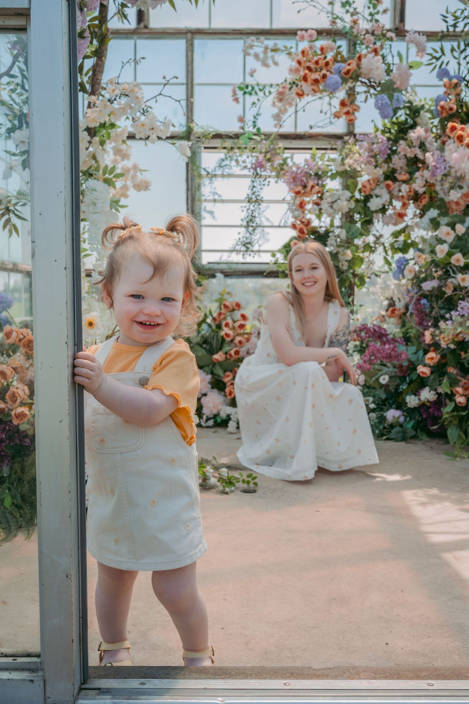 Candid greenhouse photo with toddler peeking around corner amid colorful flower displays.
