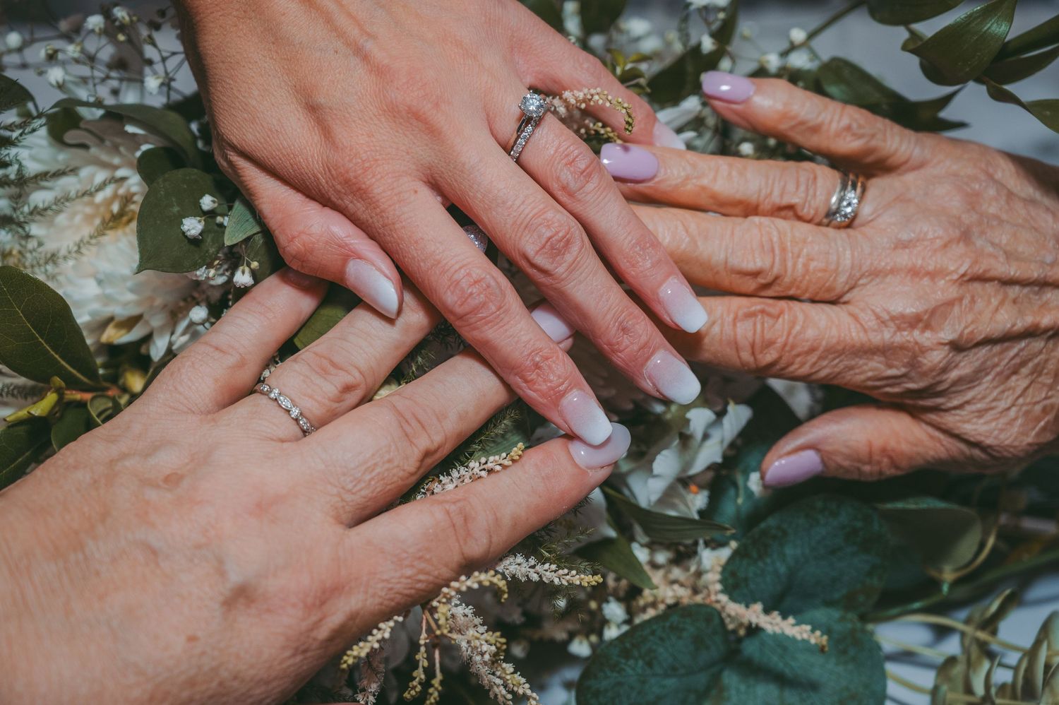 Three generations show their wedding rings in a close-up shot with floral decorations in the background.