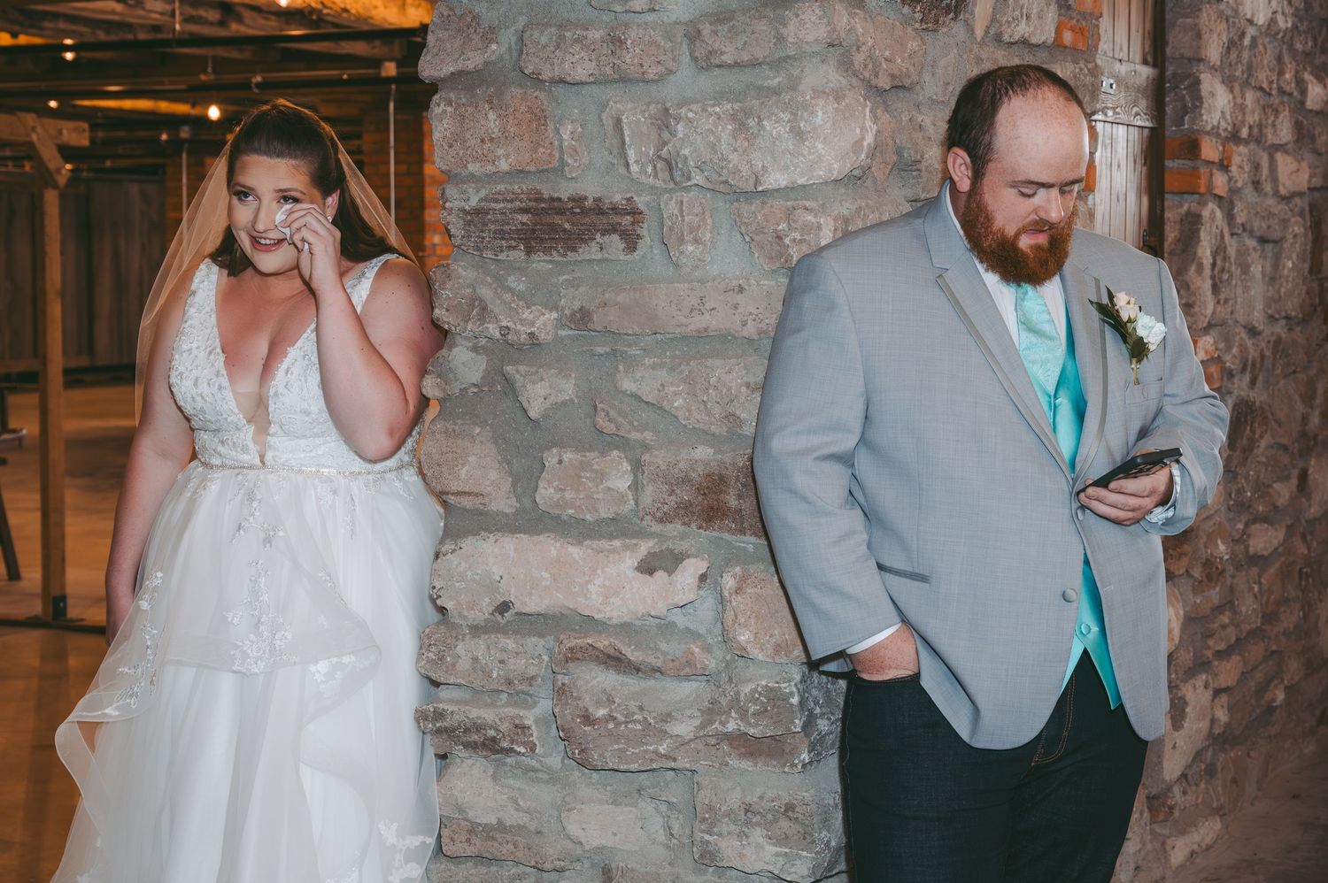 Candid moment with couple texting on phones against stone wall in wedding attire.
