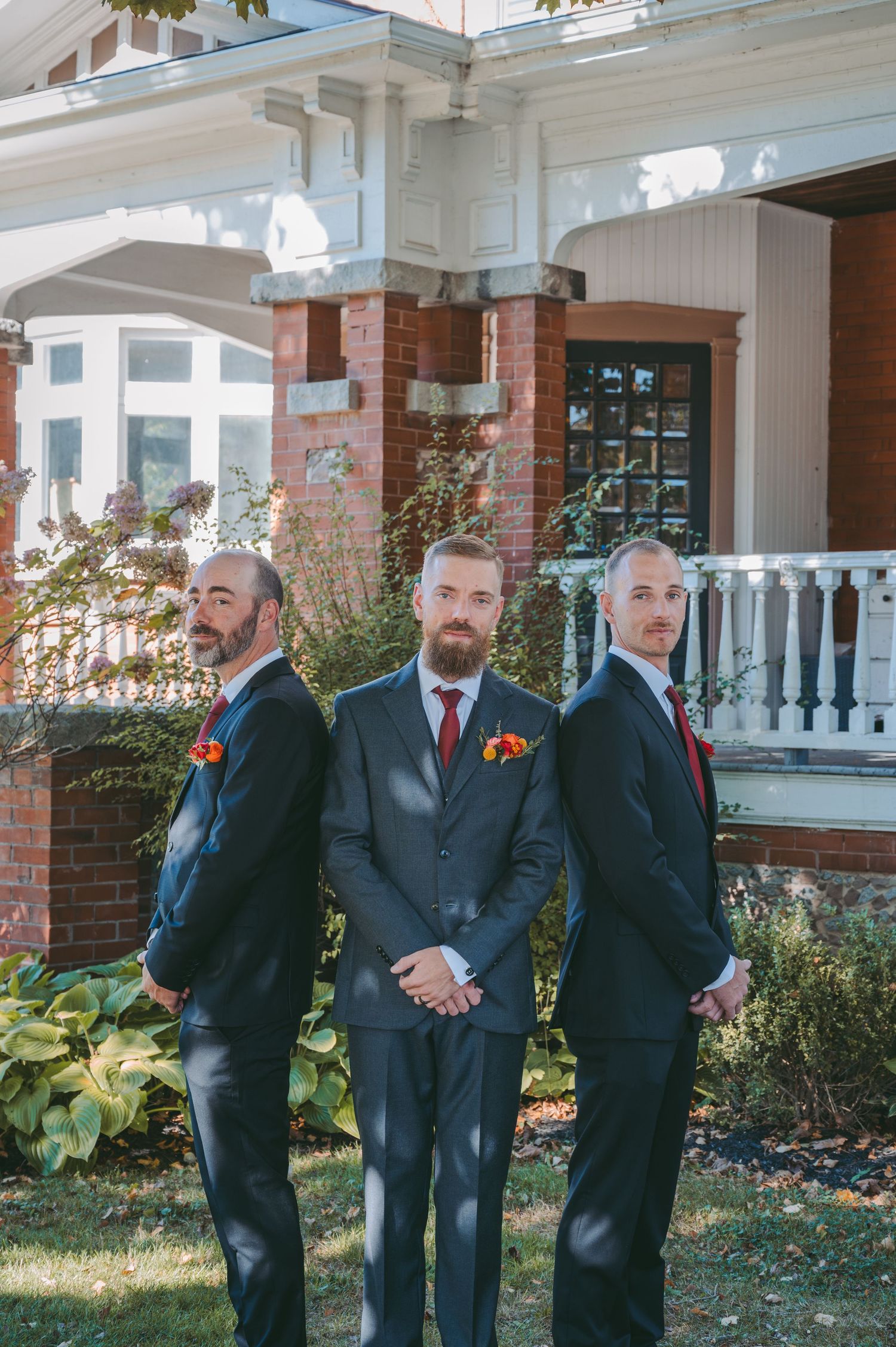 Three groomsmen in dark suits pose back-to-back with orange boutonnières outside historic venue.