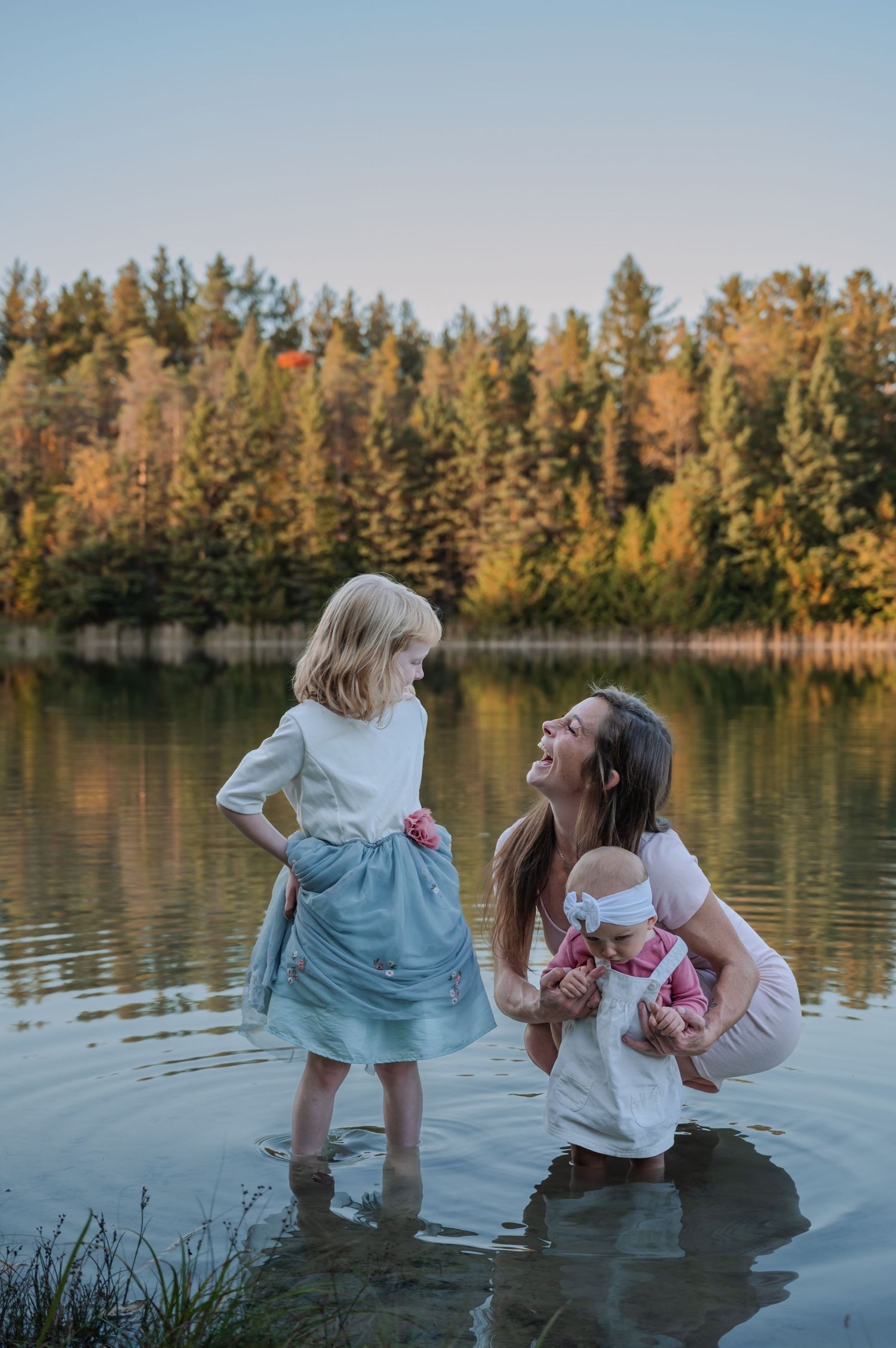 Mother and daughters wade into a serene lake at sunset with evergreen trees reflecting in the calm water.