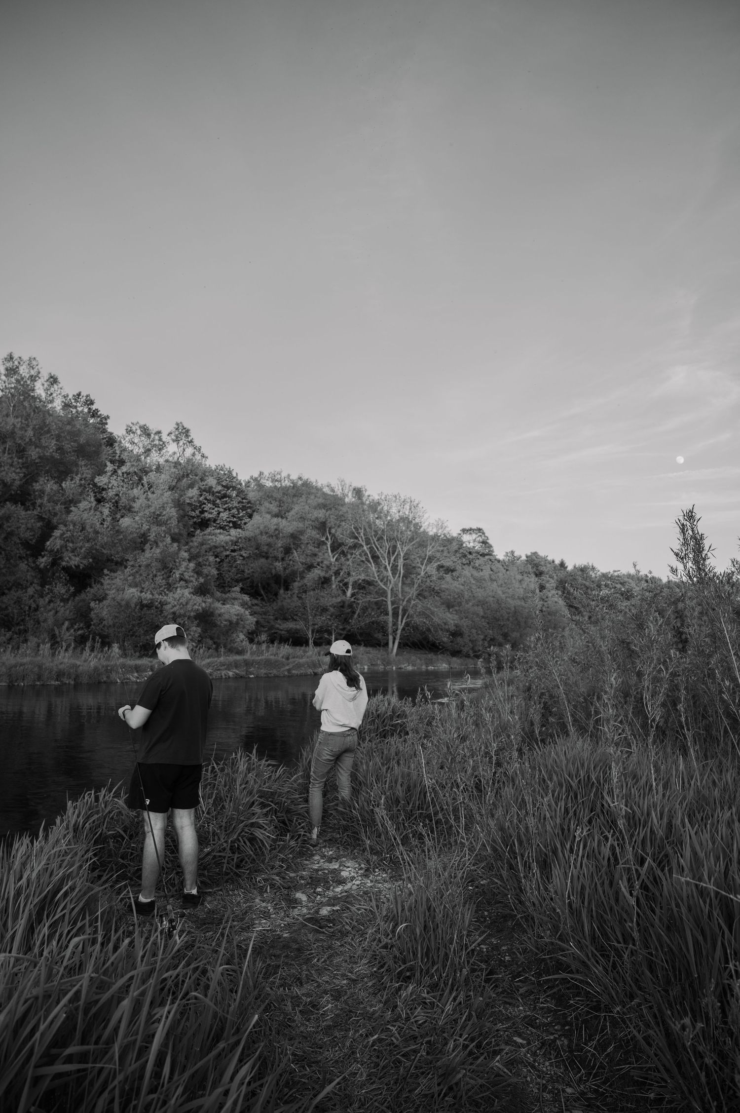 Two people fishing on the grassy bank of a serene lake surrounded by lush trees in a black and white photo.