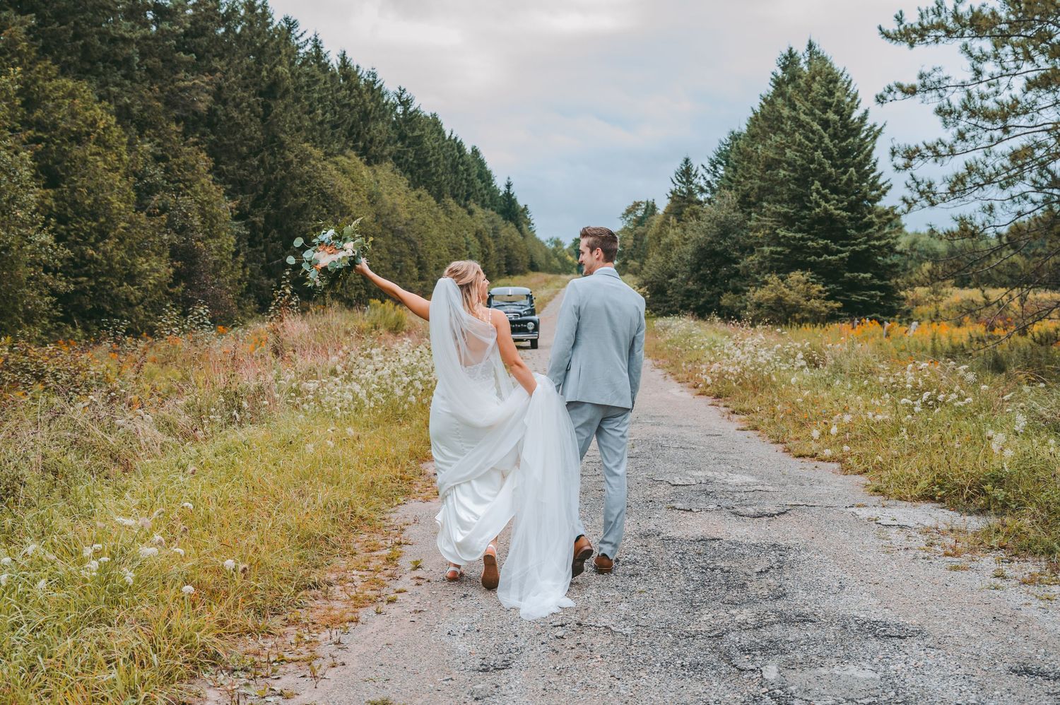 Couple walking hand in hand down a scenic mountain path surrounded by evergreen trees and wildflowers.