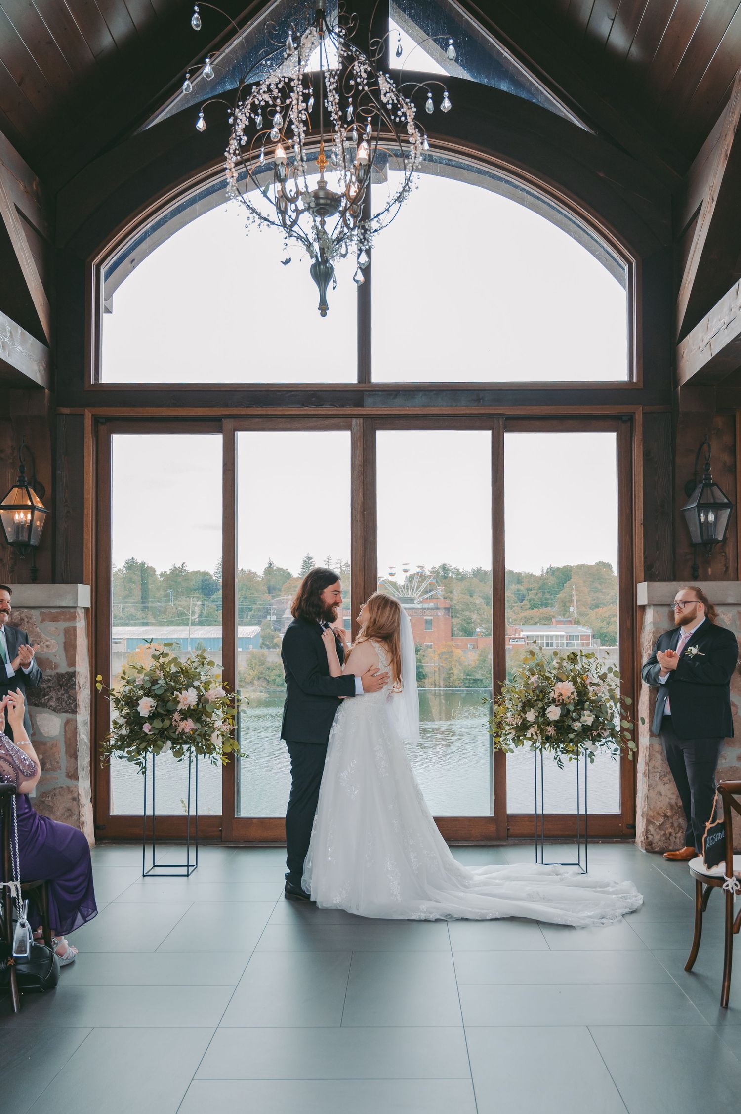 A couple shares their wedding ceremony at the Cambridge Mill with large windows, chandelier, and floral arrangements.