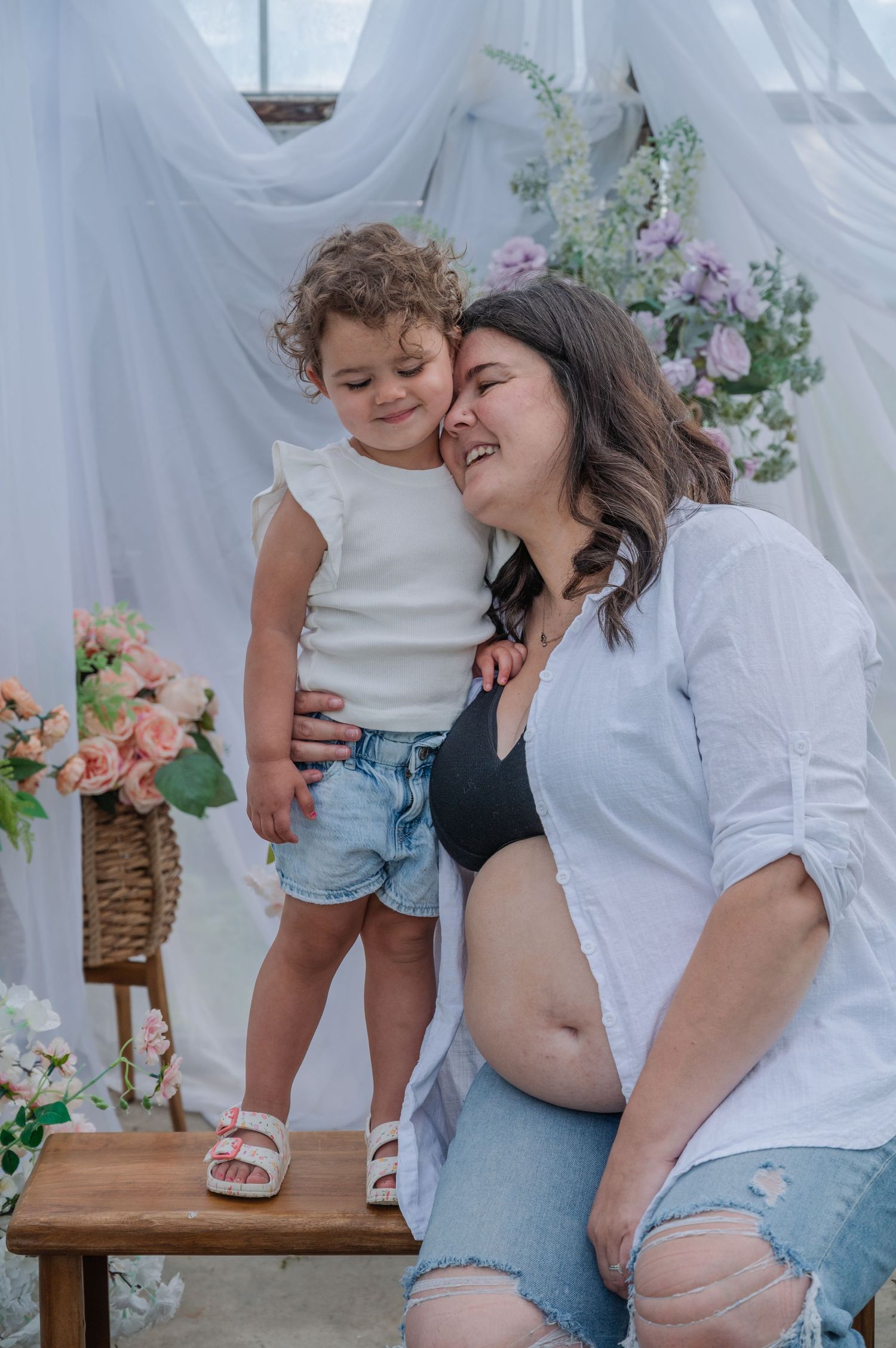 Pregnant woman sitting with toddler leaning in for a kiss during a floral maternity photo session.