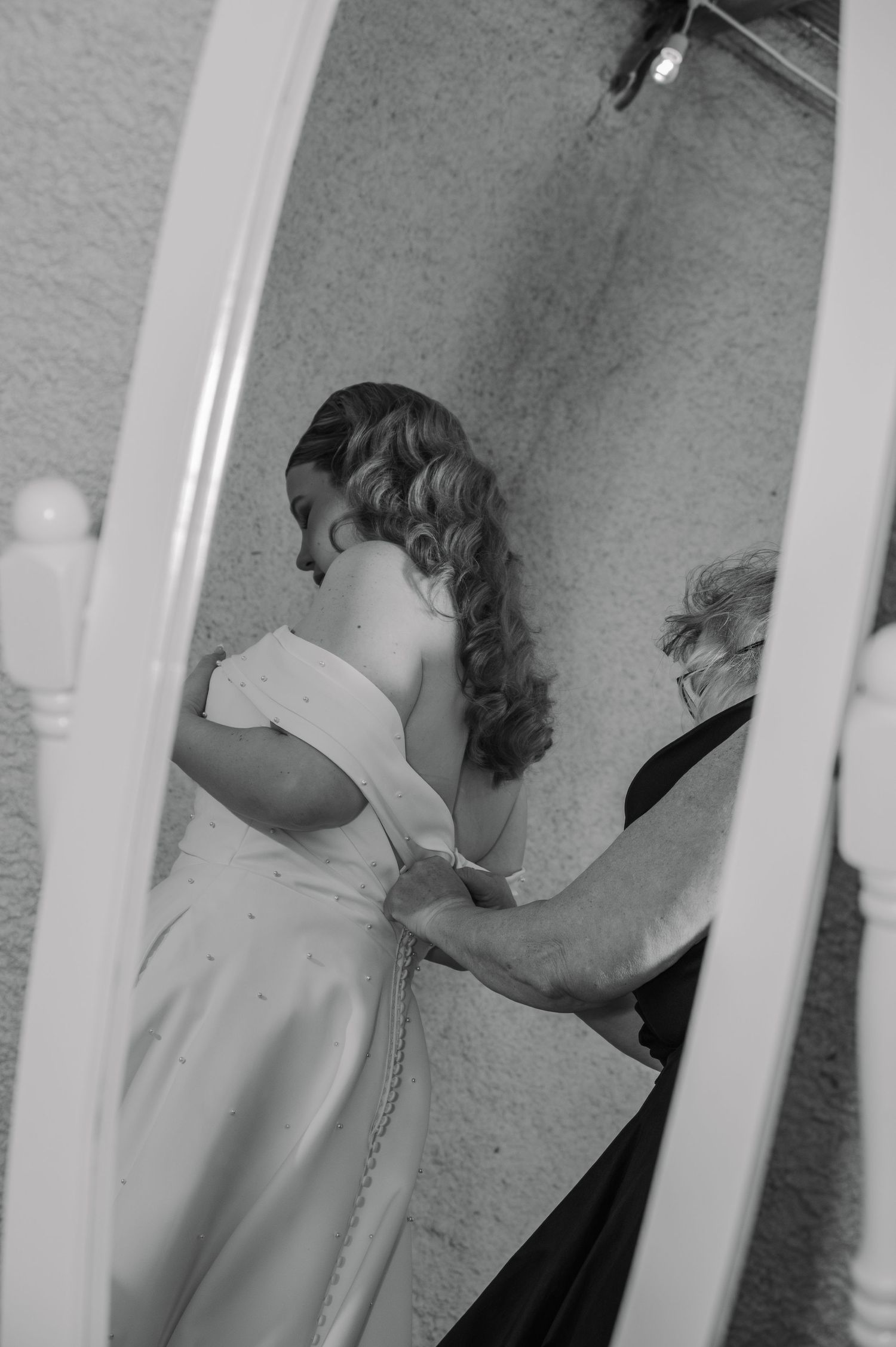Black and white photo of a bride getting her wedding dress fastened by a helper, reflected in a white framed mirror.