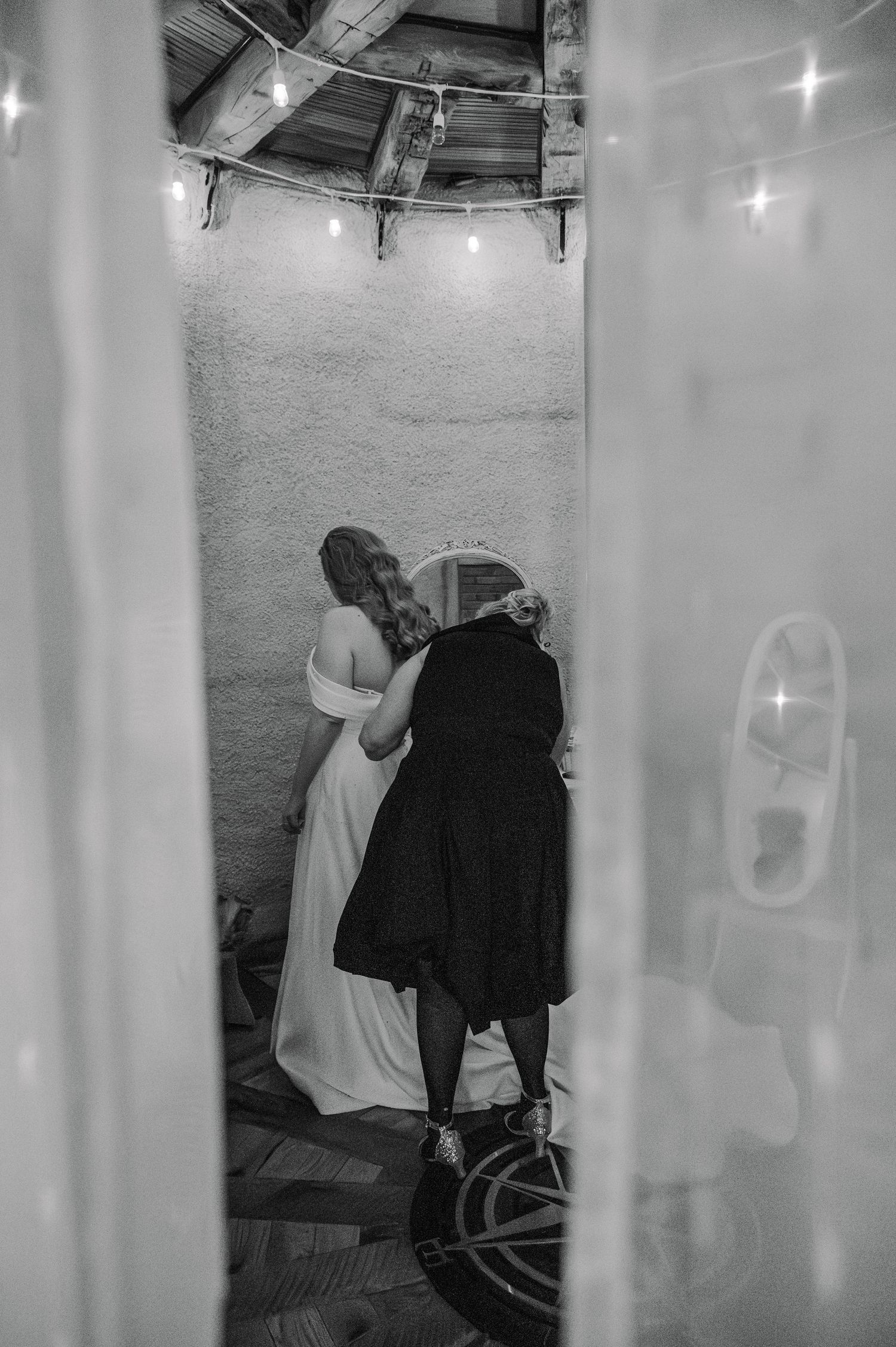 Black and white wedding photo of bride and groom embracing in a rustic venue with string lights overhead.