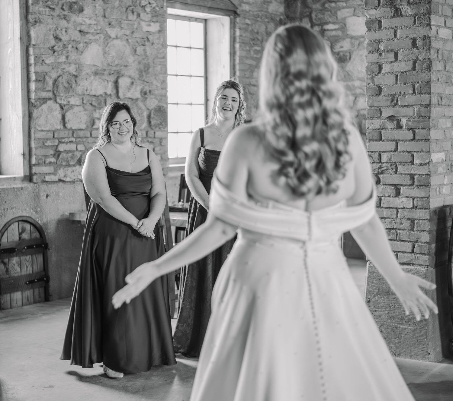 Bride in white gown reveals herself to smiling bridesmaids in dark dresses inside a rustic stone venue.