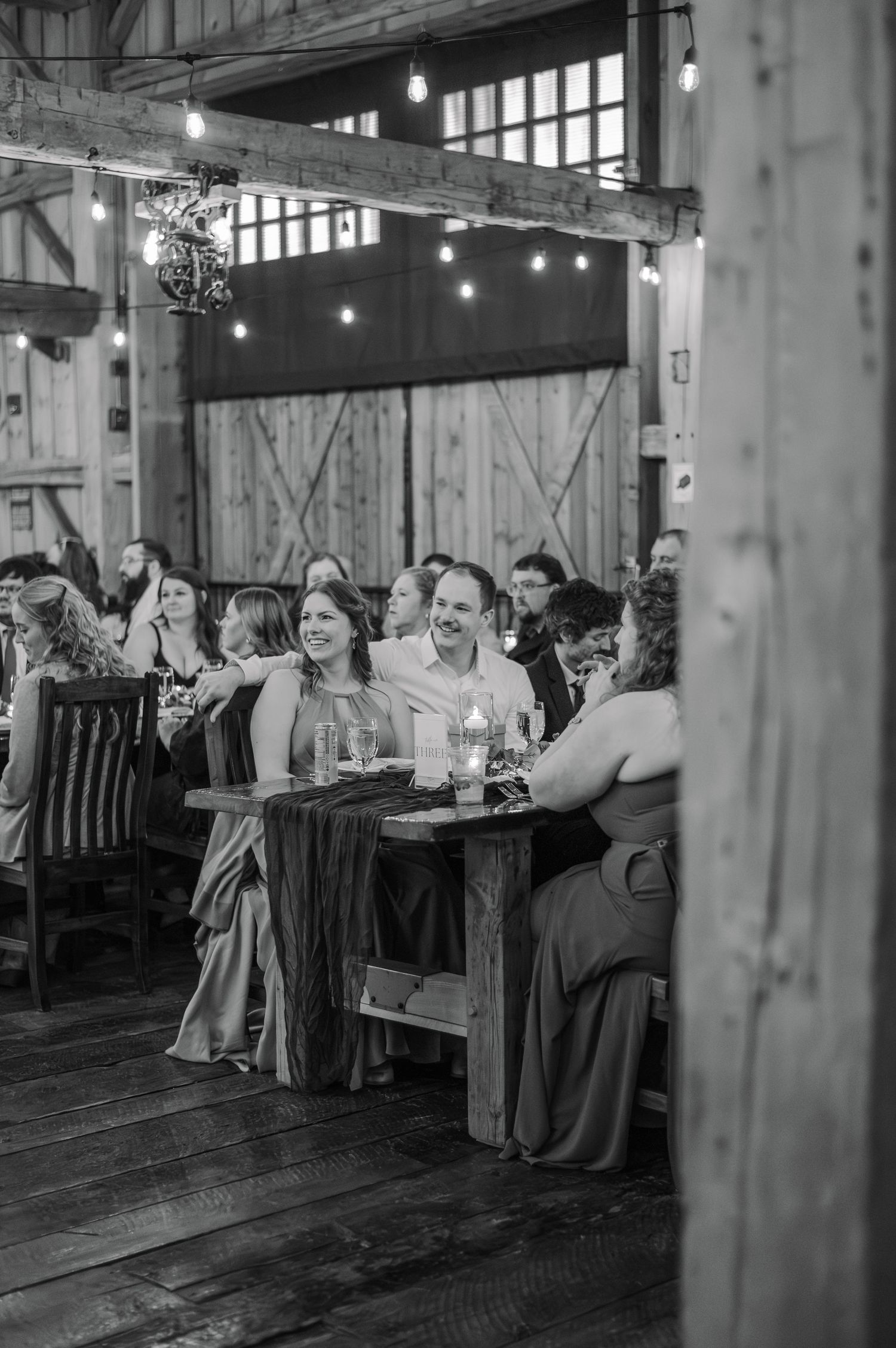 Wedding guests laugh and celebrate at rustic wooden tables inside a barn reception venue, black and white.