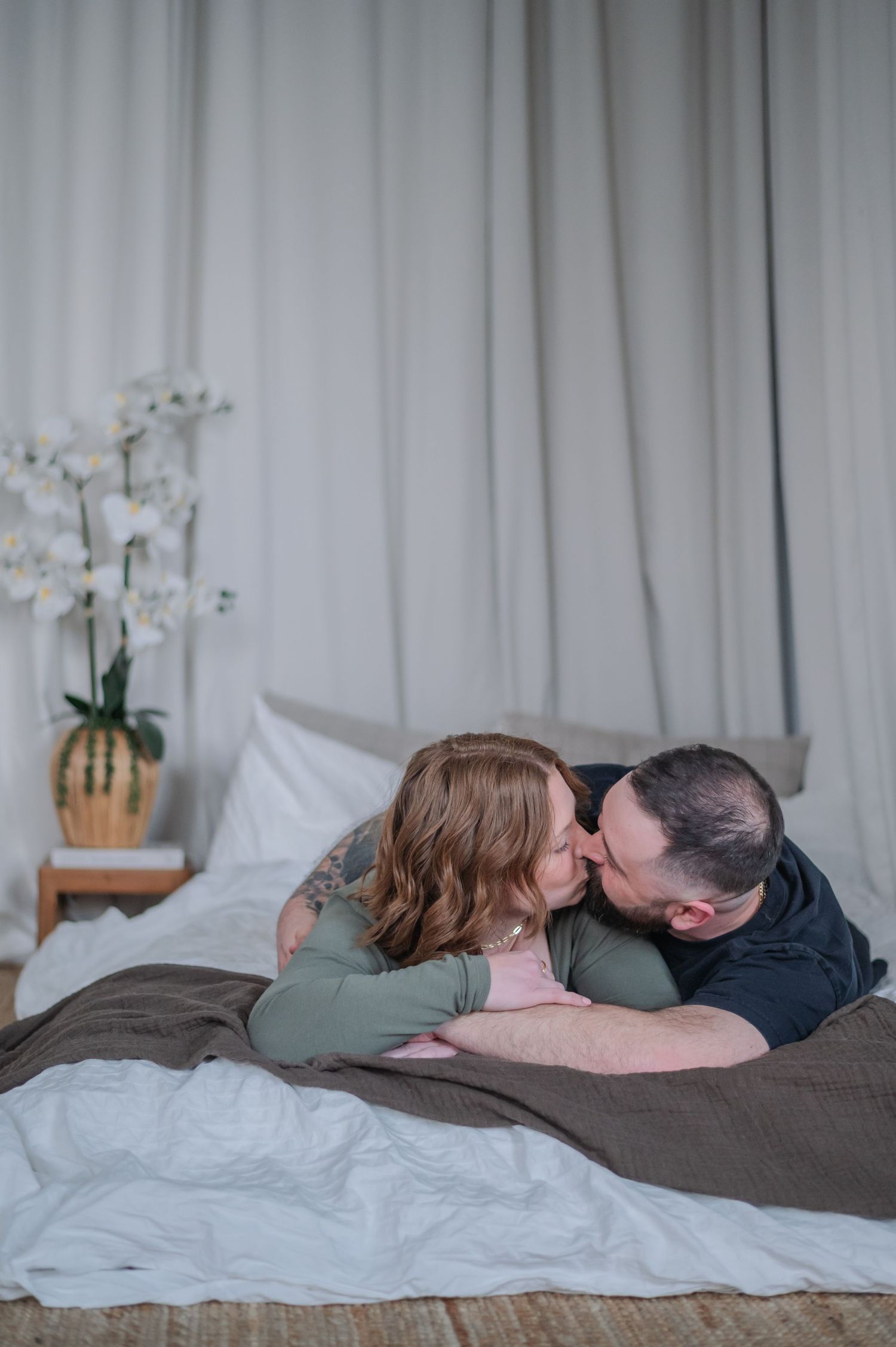 Couple sharing a tender kiss while lying on a bed with white linens and orchid flowers nearby.