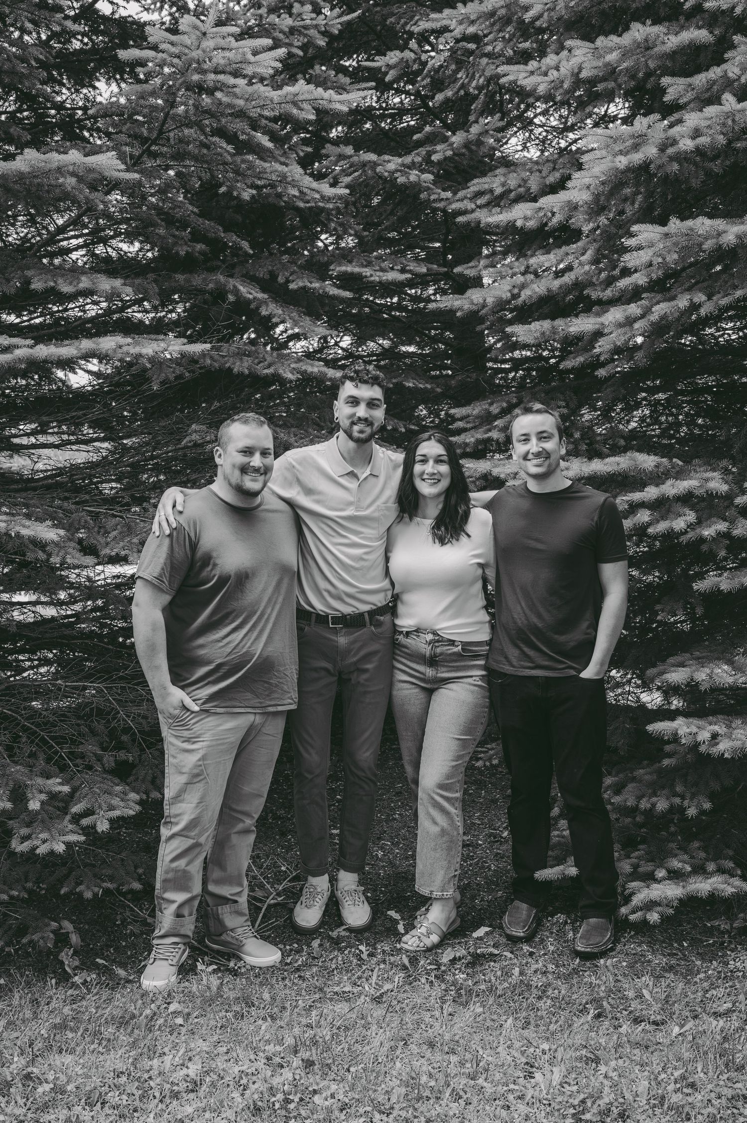 A group poses together against a backdrop of evergreen trees in a black and white photograph.