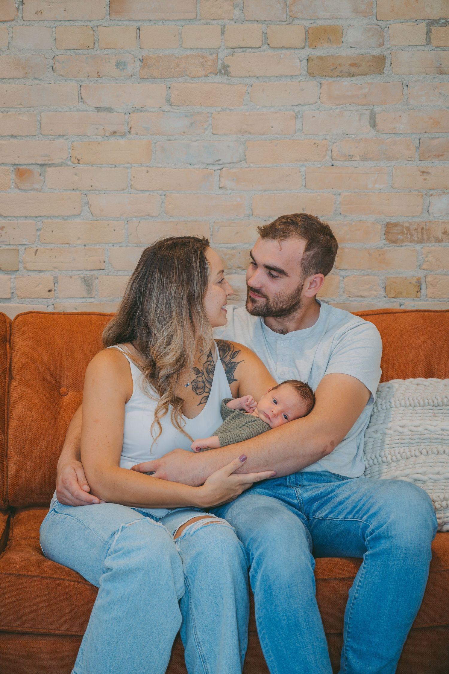 Family of three snuggling on orange couch against exposed brick wall.