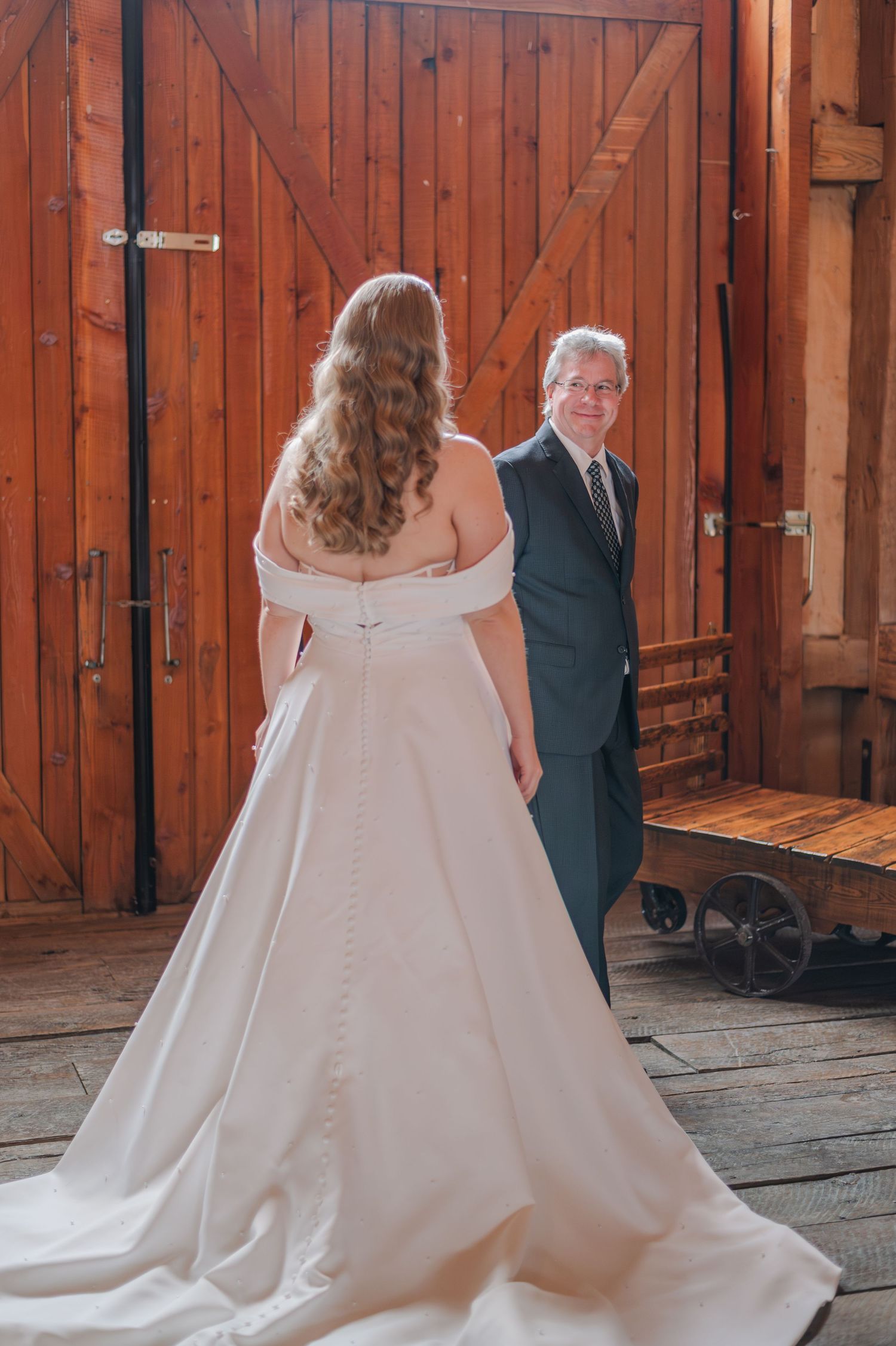 Bride in off-shoulder white gown with train faces groom in dark suit near rustic wooden barn doors.