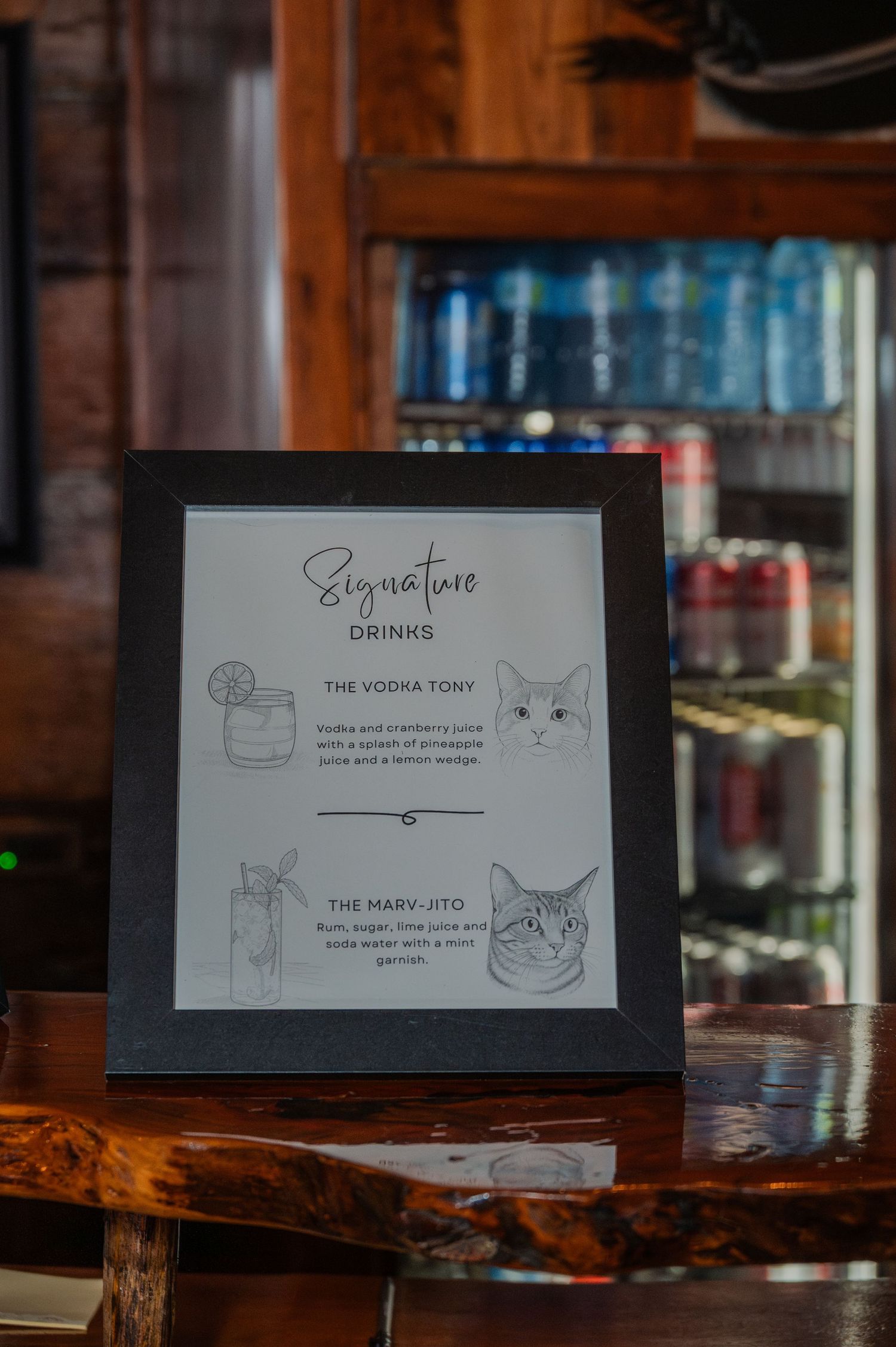 Framed signature drinks menu featuring cat illustrations for The Vodka Tony and The Marv-Jito cocktails at a bar.